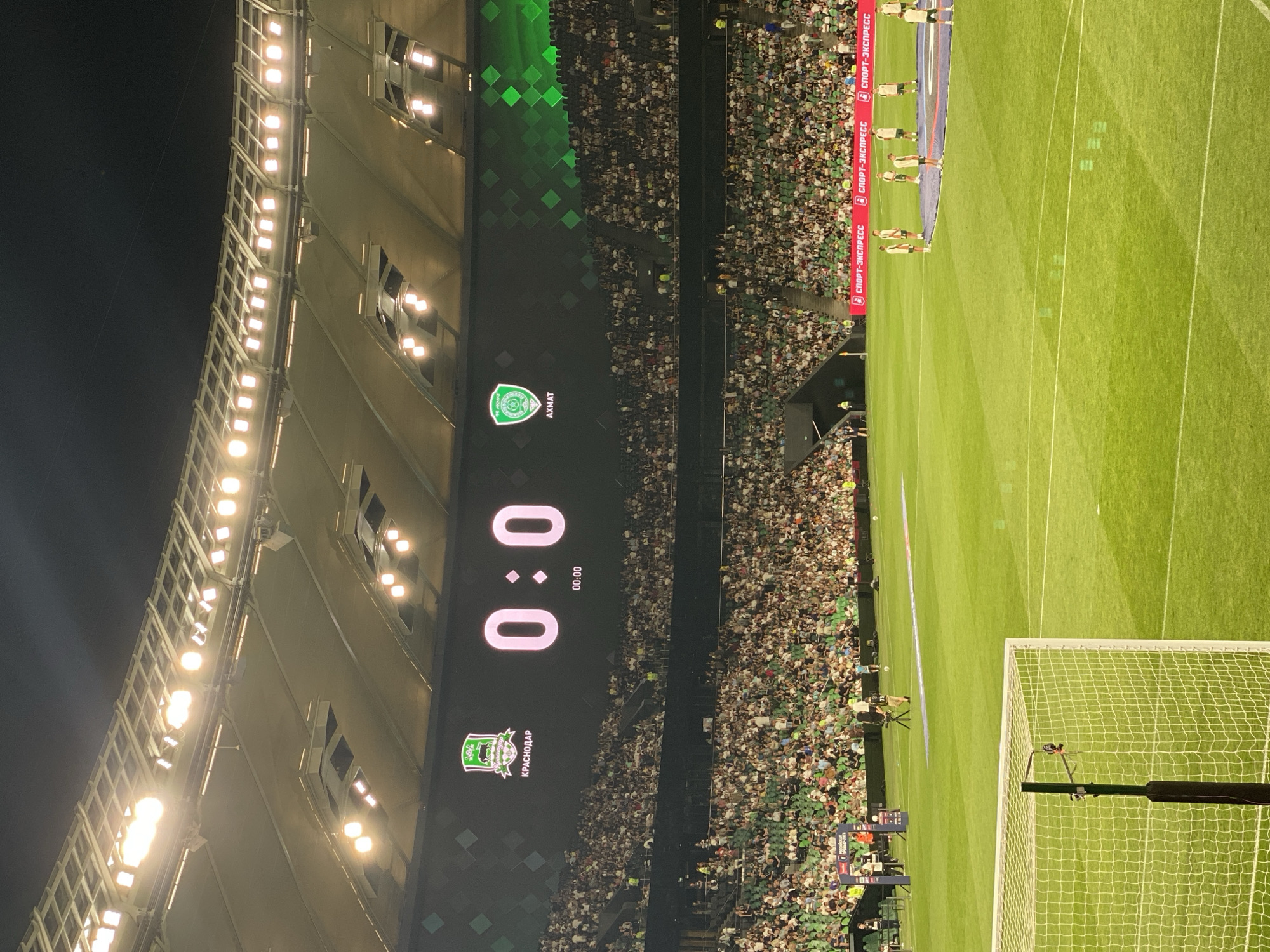 A wide-angle, nighttime photograph captures a soccer stadium filled with spectators. A large scoreboard in the background displays the score "0:0" with a timer showing "00:00." Two team logos are visible on either side of the scoreboard. The stadium is illuminated by bright overhead lights. The field in the foreground is green grass, marked with white lines. Several players in uniform are standing on the field. The stands are densely populated with a crowd wearing various clothing. There are cameras and other equipment set up around the perimeter of the field.