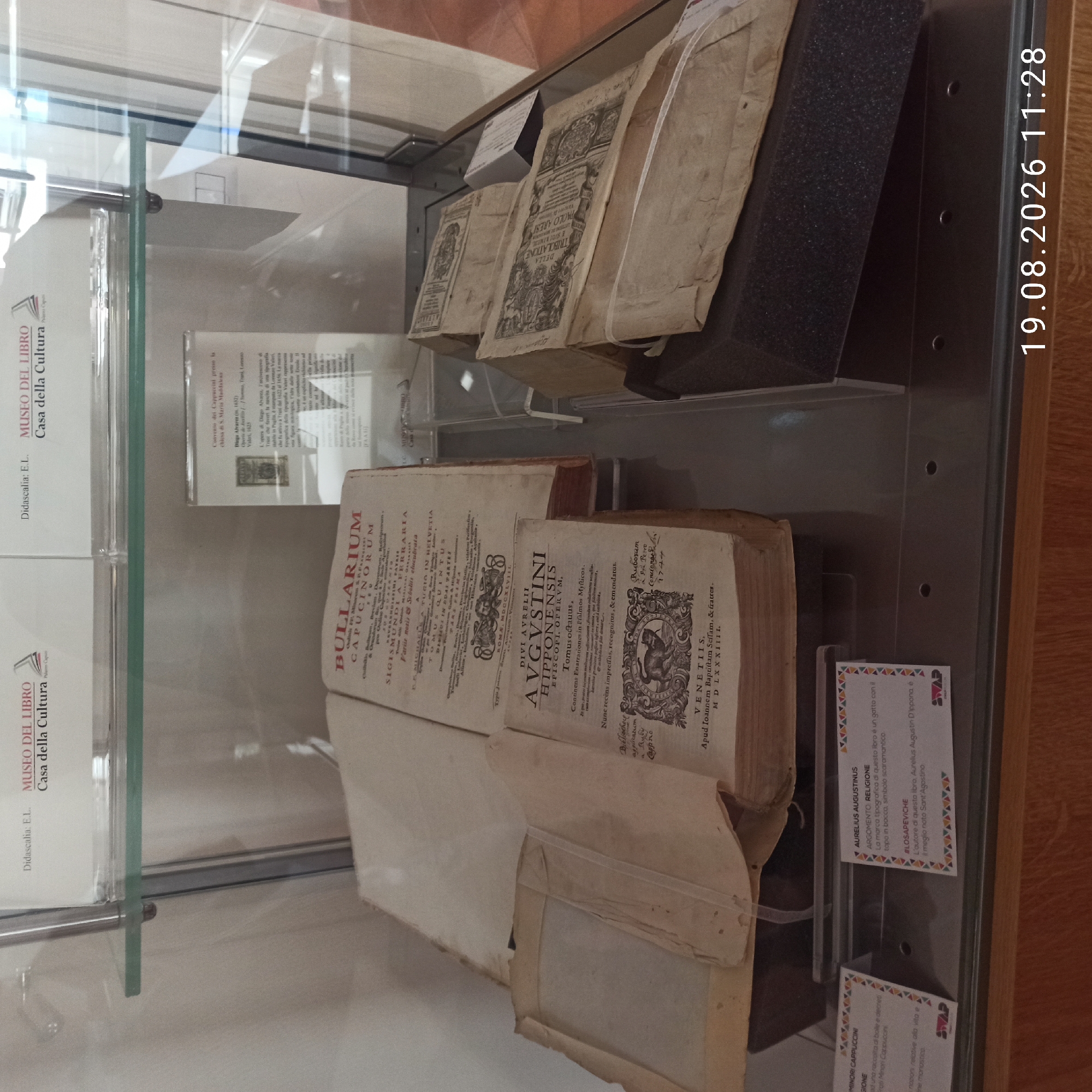 The image shows a glass display case containing antique books and documents, likely in a museum or library. On the top shelf, two framed items with logos of "MUSEO DEL LIBRO" and "Casa della Cultura" are visible. Below, on the main shelf, several bound volumes are displayed. One prominent open book has the title "BULLARIUM CAPUCINORUM" on its cover. To its right, another open book displays "DIVI AVGVSTINI HIPONENSIS EPISCOPI OPERVM". Several other stacked and wrapped books are also present. Small descriptive labels are attached to the front of some items. The overall environment appears to be indoors, with artificial lighting. The timestamp "19.08.2026 11:28" is superimposed on the bottom right of the image. The scene is devoid of people. The location is identified by the presence of texts mentioning "MUSEO DEL LIBRO Casa della Cultura". Although the specific city of Ruvo di Puglia is not directly visible in the image, such a museum would be a point of cultural interest within that city.