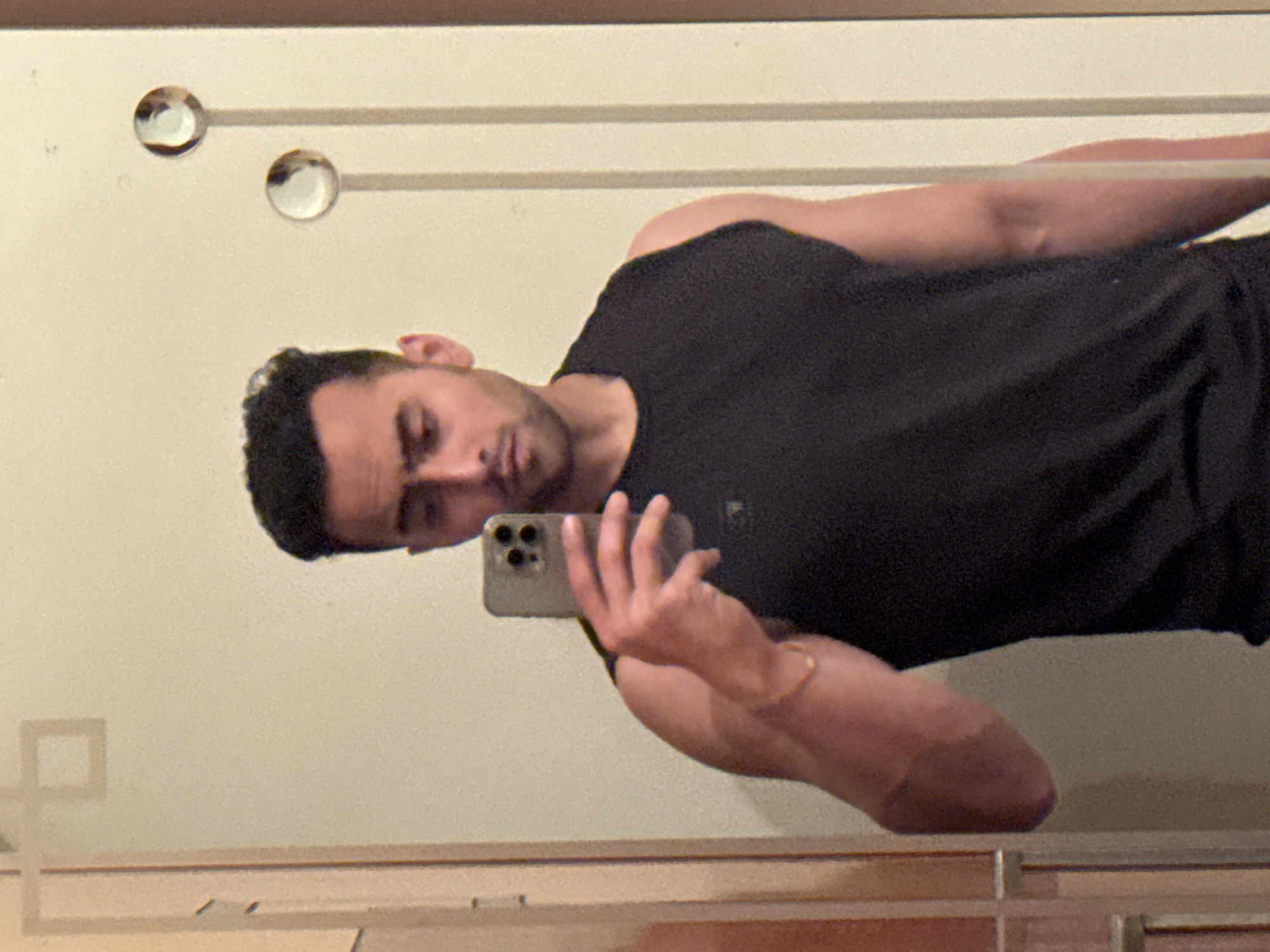 A man is taking a selfie in front of a mirror. He is holding a smartphone in his right hand. He has dark hair and is wearing a black t-shirt. The background is a light-colored wall with decorative elements. The location is difficult to ascertain from the image alone, and there is no definitive evidence indicating Kolkata, India.