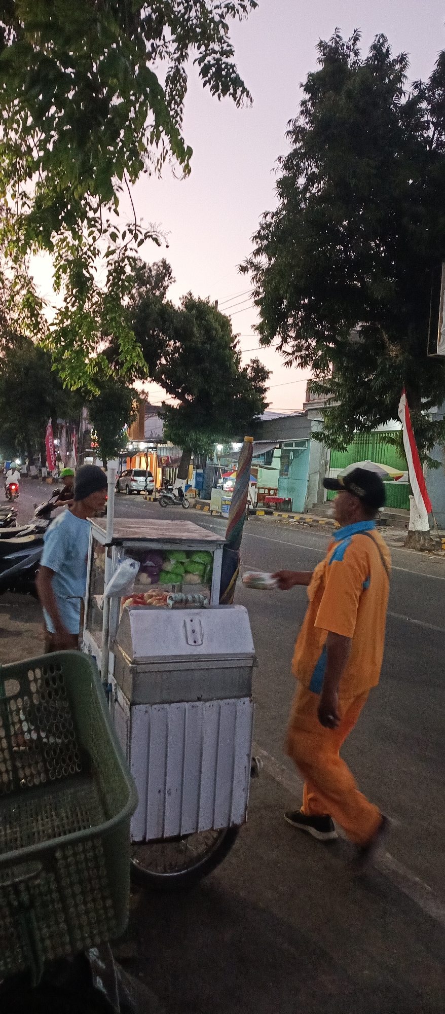 In Ardirejo, Indonesia, at dusk, a street vendor pushes a food cart on the side of a road. The cart is white with a glass display case on top, filled with food items. Two men are interacting near the cart. One man, wearing a light blue shirt, stands by the cart. The other man, wearing an orange shirt and pants and a black cap, is reaching out to the cart, seemingly receiving an item. The road is paved, with a white line marking the lane. Trees line the side of the road, and buildings are visible in the background, illuminated by the fading daylight. Several motorbikes are parked or passing by. A green plastic basket is positioned in the foreground. A flag, red and white, is visible on a pole to the right. The sky is a soft gradient of pink and purple.