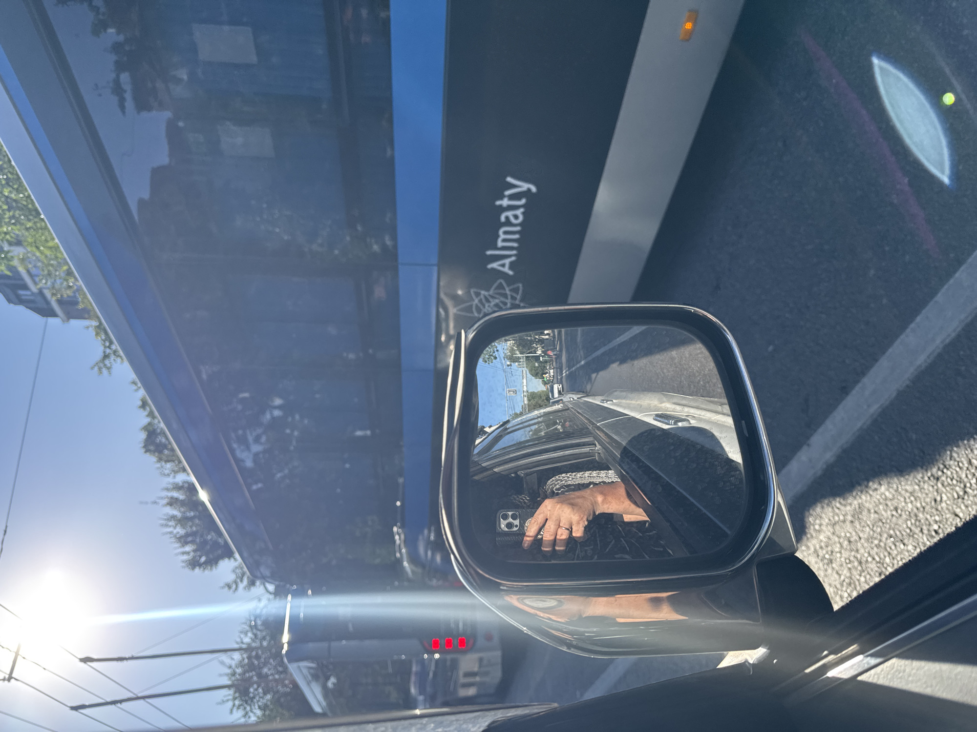 The image shows a view from inside a car, looking out of the side window and into the side-view mirror. The side-view mirror reflects a person's hand holding a smartphone, taking a selfie. The person is wearing a ring and a bracelet. The car appears to be in traffic, as evidenced by the bus in front and the cars visible in the mirror's reflection. The bus has the word "Almaty" written on its side, indicating the location is Almaty, Kazakhstan. The sun is shining brightly, creating lens flare and casting shadows. The environment appears to be an urban street with trees and buildings visible in the background.