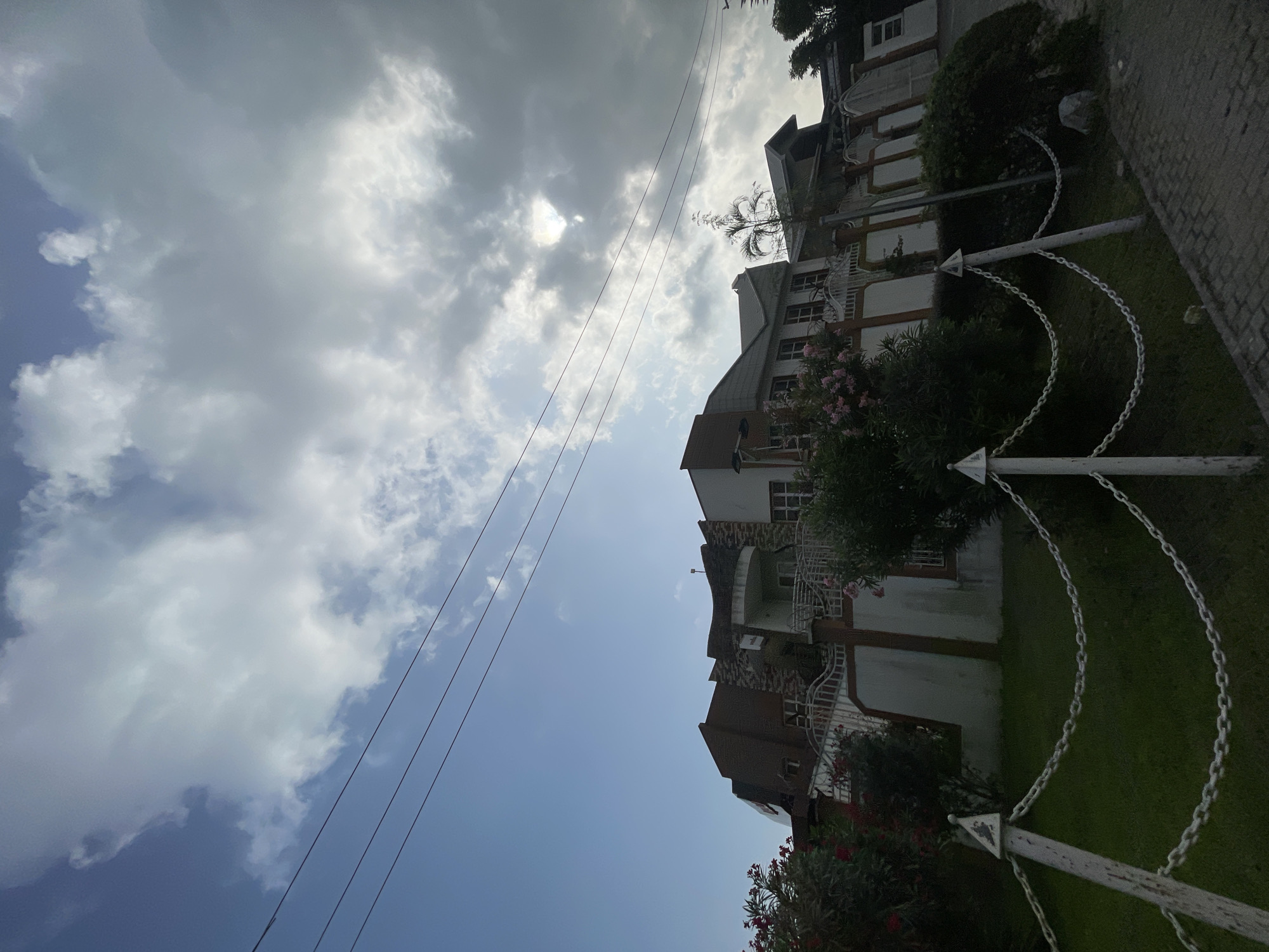 The image depicts a multi-story building in Moba, Nigeria, set against a sky with partly cloudy conditions. Power lines run across the sky. The building appears to have multiple balconies and is bordered by greenery, including trees and flowering shrubs. A white chain-link fence is present in the foreground. The architecture suggests a residential or commercial structure, possibly an apartment complex or office building. The picture is taken from ground level looking up at a slight angle.