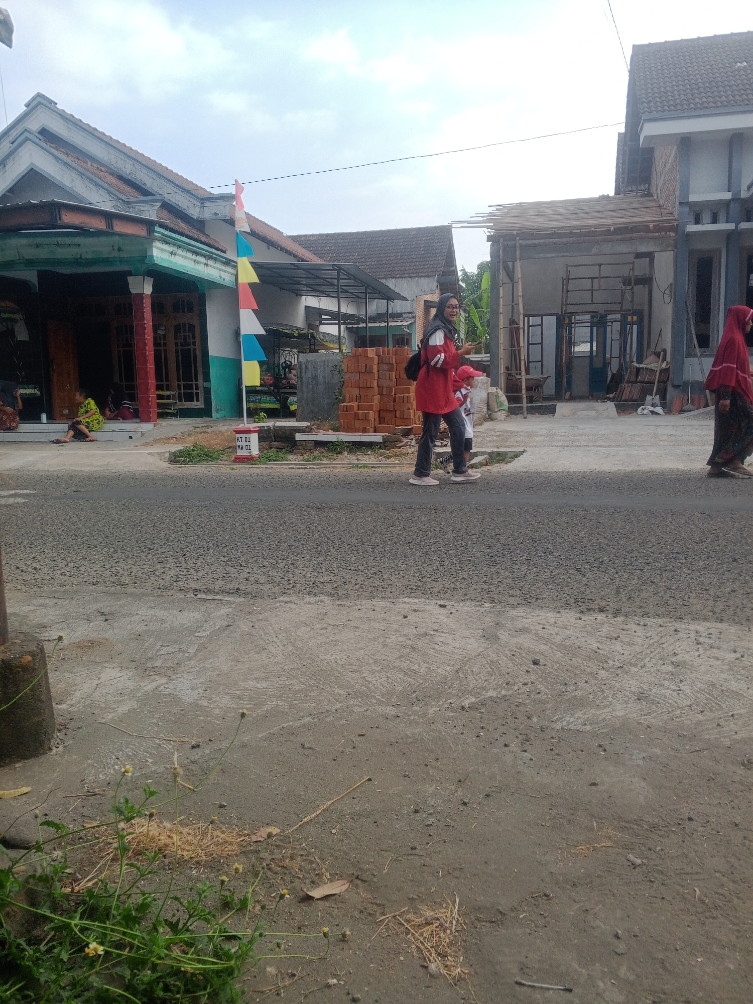 The image depicts a street scene in Lembah, Indonesia. In the foreground, a gray asphalt road is visible, with patches of concrete and dry grass. A concrete post stands on the left side of the road. In the midground, a few individuals are present. On the left, two people are sitting on a porch of a building, one wearing a green and yellow patterned dress and the other in red. A sign with "RT 01 RW 01" is displayed near the building. Further to the right, a woman in a red jacket and hijab, and a child in a red hat and white shirt are walking on the sidewalk. Another person in a red hijab and dark patterned clothing is walking further down the street to the right. Behind them, a building under construction is visible with scaffolding and construction materials like bricks. The background features more residential buildings with tiled roofs and a cloudy sky.