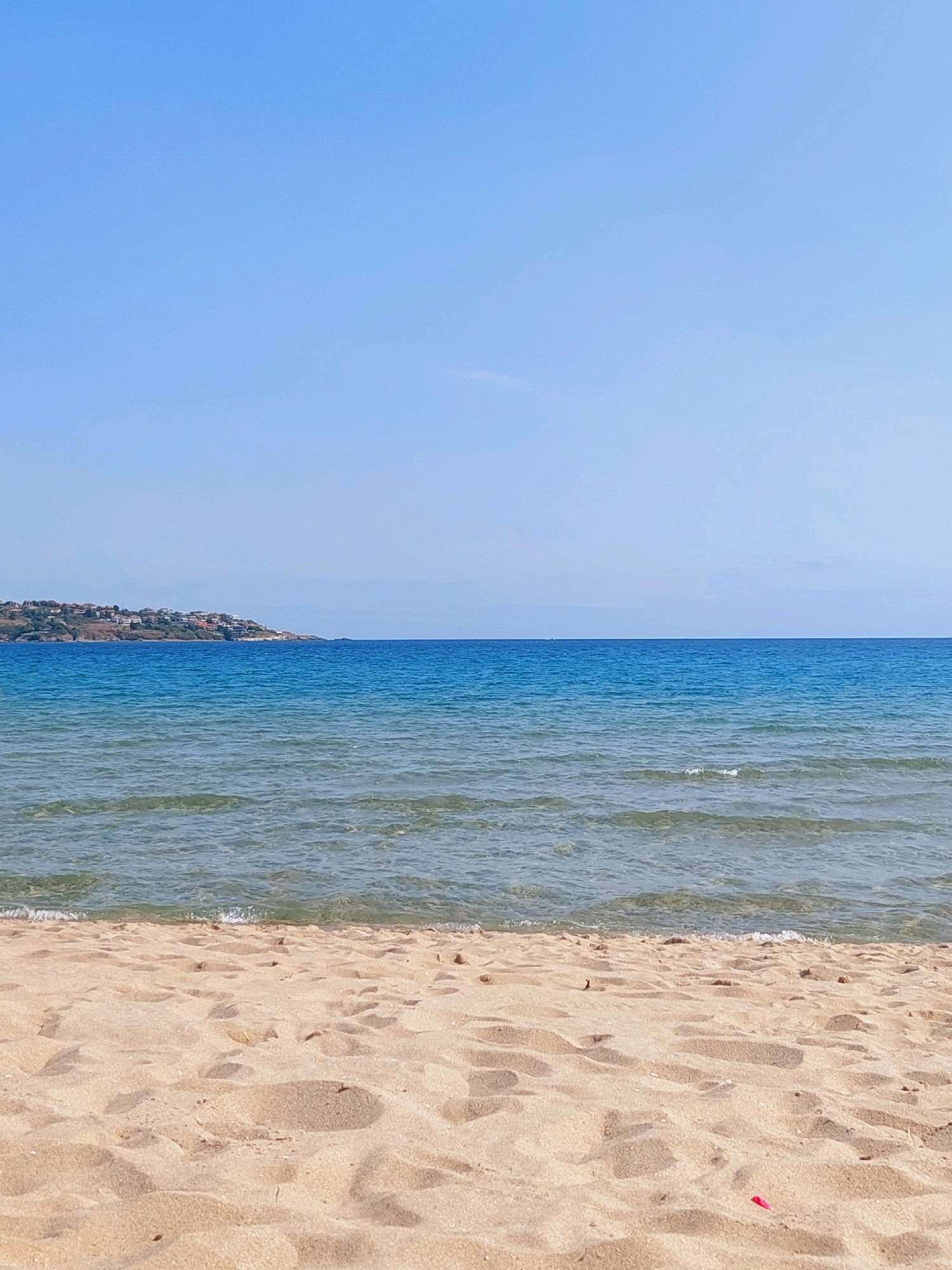 The image depicts a serene beach scene with a clear blue sky dominating the upper half. The horizon line, where the sky meets the sea, is sharp and distinct. The ocean stretches out, transitioning from a lighter, translucent blue near the shore to a deeper, richer blue further out. Gentle waves are visible, with small whitecaps indicating movement. In the distance, a densely populated coastline with buildings is visible on a gently sloping hill. The foreground is occupied by a sandy beach, showing the texture of the sand with subtle variations in color and light, indicating it is dry and undisturbed. A small red object is visible on the sand in the lower right corner. This scene is characteristic of the beaches found in and around Sozopol, Bulgaria, a historic coastal town known for its picturesque seaside.