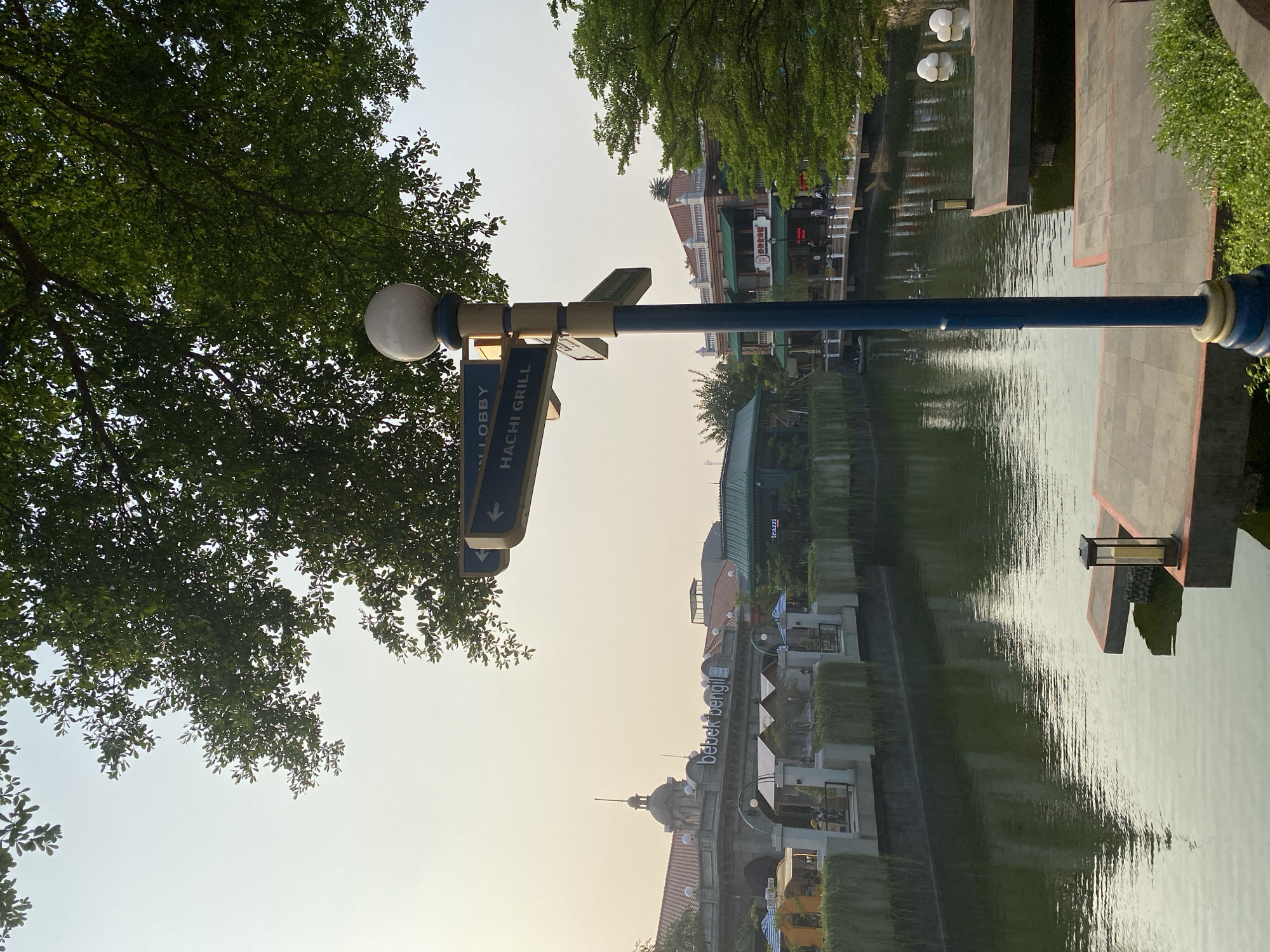 The image depicts a canal-side promenade with buildings and lush greenery. A prominent blue signpost with white lettering, pointing to "LOBBY" and "HACHI GRILL," stands in the foreground, partially obscured by the branches of a large tree. The canal reflects the architecture and the sky, suggesting a serene atmosphere.
Buildings with varied architectural styles line the canal. One building displays the signage "bebek bengil," an Indonesian phrase likely indicating a restaurant. Other structures appear to be commercial establishments or residences, with balconies and outdoor seating areas visible. The presence of umbrellas suggests outdoor dining or a sunny day.
In the distance, people can be seen strolling along the promenade and in the cafes, indicating a leisurely activity or event. The time of day appears to be late afternoon or early evening, with a soft, diffused light and the sky showing a pale hue. The weather seems pleasant, conducive to outdoor activities.
The overall scene conveys a sense of tranquility and a pleasant urban environment, blending natural elements like trees and water with commercial and residential architecture. The reflection on the water adds a dynamic visual element to the static structures. The location, East Jakarta, Indonesia, hints at a tropical climate and a vibrant urban setting.