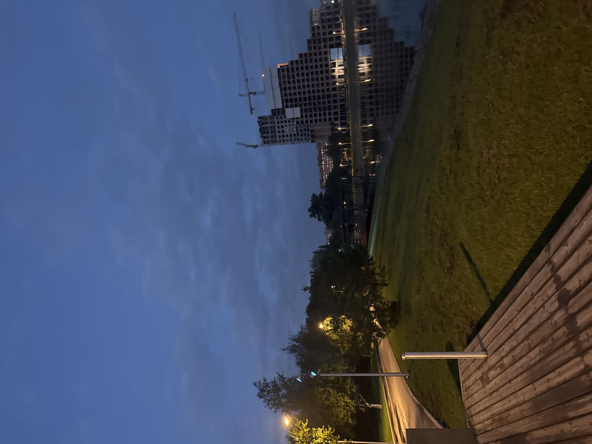 The image depicts a twilight scene by a body of water, likely a river or lake, in Ufa, Russia. A large, partially constructed building dominates the right side of the frame, with several cranes visible against the darkening sky, suggesting ongoing construction. The building's reflection is visible in the calm water.
To the left, a pathway curves through a grassy area with trees. Streetlights illuminate parts of the pathway and the foliage, casting long shadows on the grass. In the foreground, a wooden deck or boardwalk is visible, its planks showing signs of wear. The sky is a deep blue, with some clouds present. The overall atmosphere is serene, with the lighting indicating either early morning or late evening. No people are visible in the image.