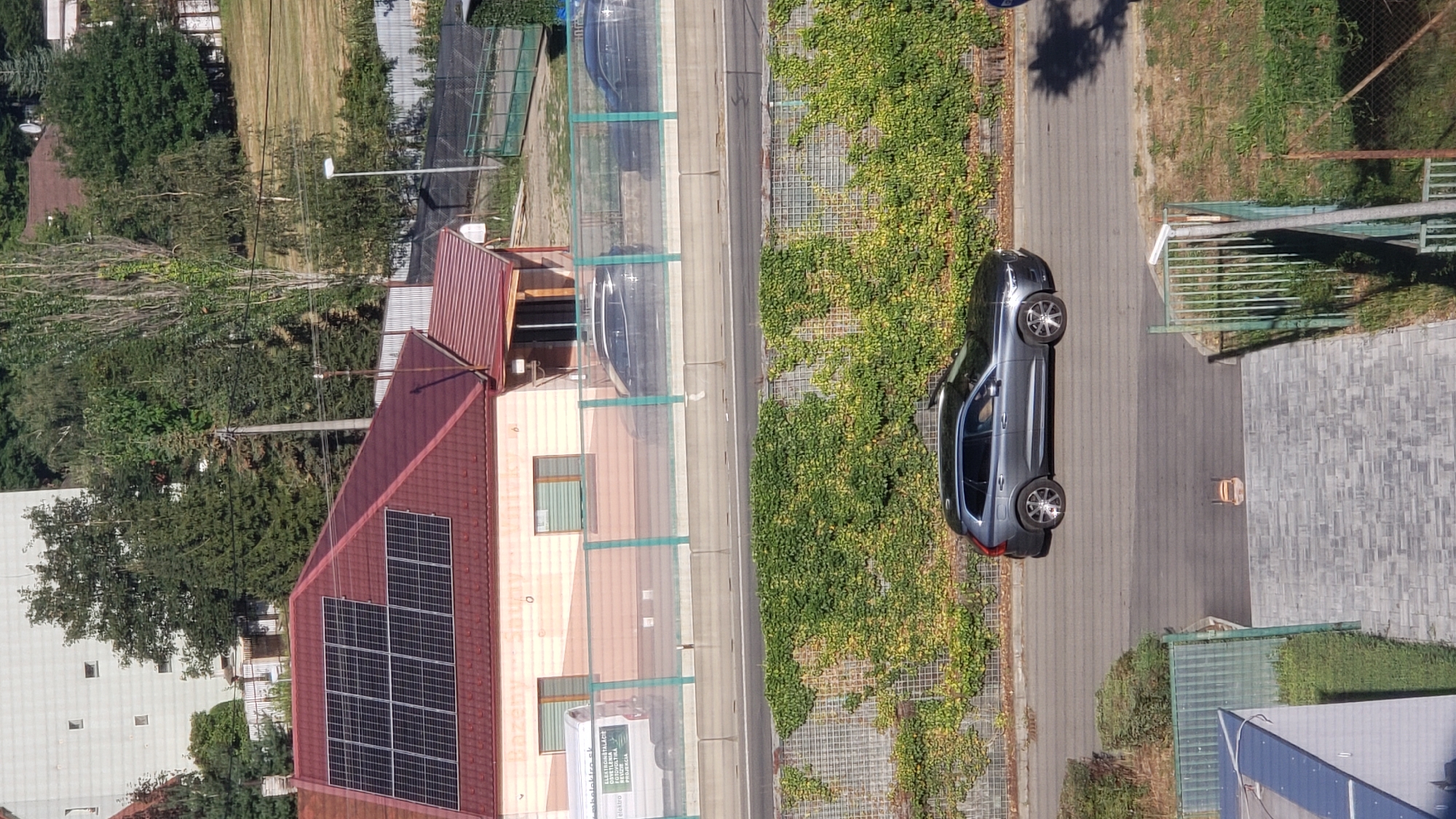In Prešov, Slovakia, an elevated perspective captures a street scene dominated by residential and commercial buildings, a highway, and lush greenery. A grey Volvo SUV is parked on the street, its wheels visible. Behind it, a highway noise barrier is adorned with climbing plants, forming a green wall. In the background, a building with solar panels on its roof is partially visible. The overall scene suggests a suburban or semi-urban environment with a blend of infrastructure and nature.