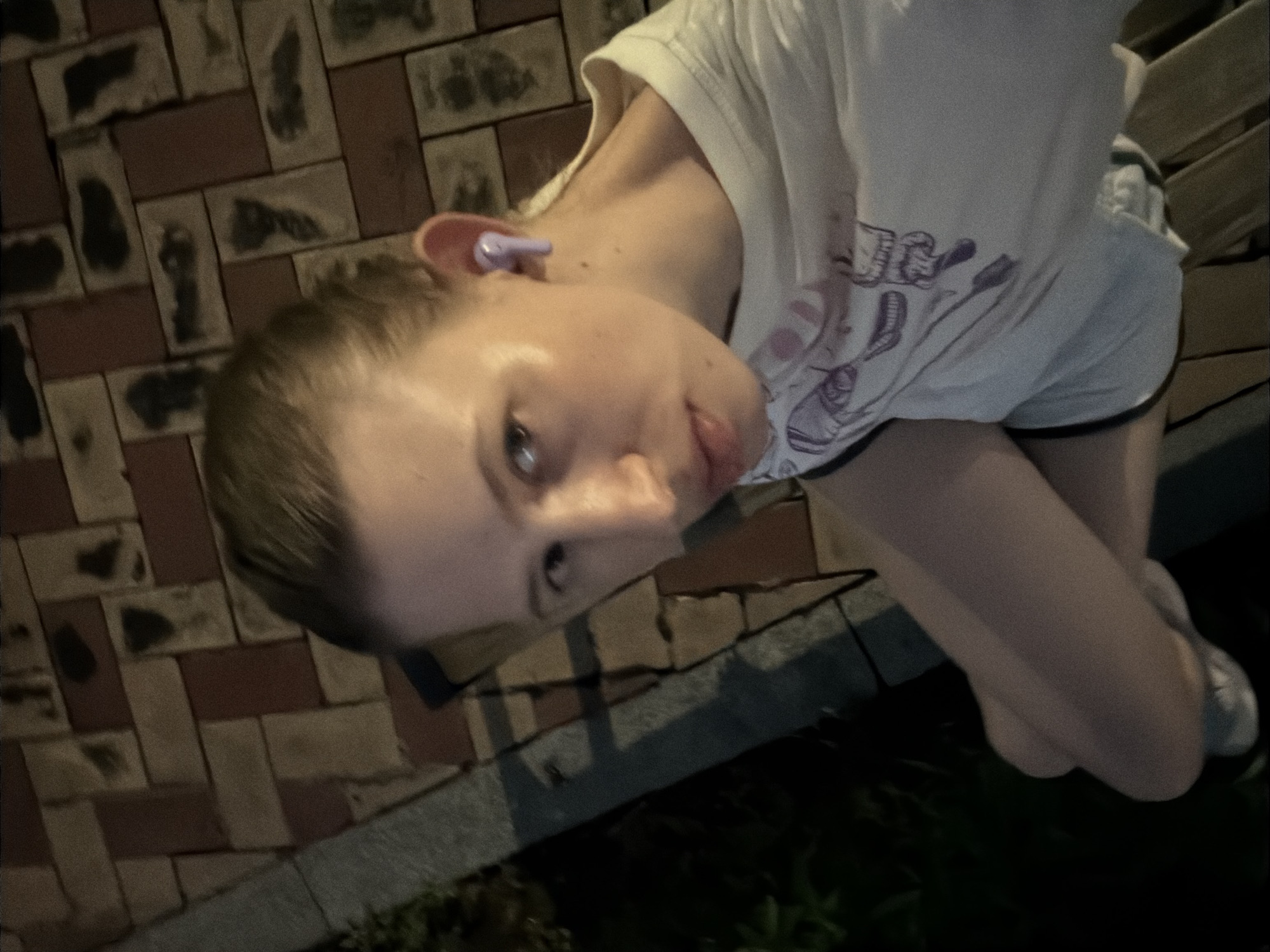 The image shows a young person sitting on a bench outdoors at night. The person is wearing a white t-shirt with a graphic design and a purple earbud in their right ear. The background consists of a tiled walkway and dark foliage. The setting appears to be a public park or pedestrian area within Dongducheon-si, South Korea. The lighting suggests artificial illumination from streetlights or nearby buildings. The person is looking towards the camera.