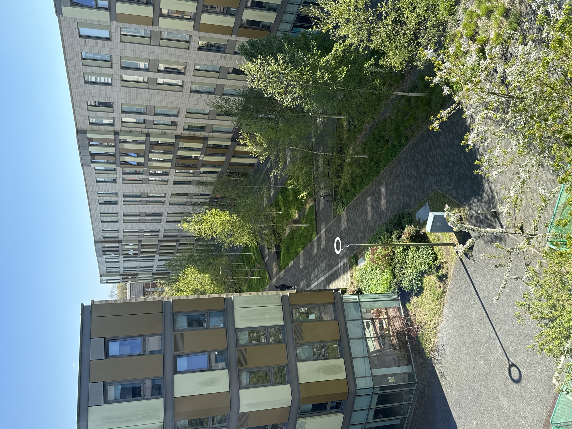 An elevated view shows a pathway running between two modern buildings, with landscaping and trees lining the path. The building to the left has a facade of beige and grey panels, while the building on the right is taller with a grey facade. A person is lying on the path between the buildings, with lampposts spaced along the pathway. This image is likely taken in Hull, United Kingdom.