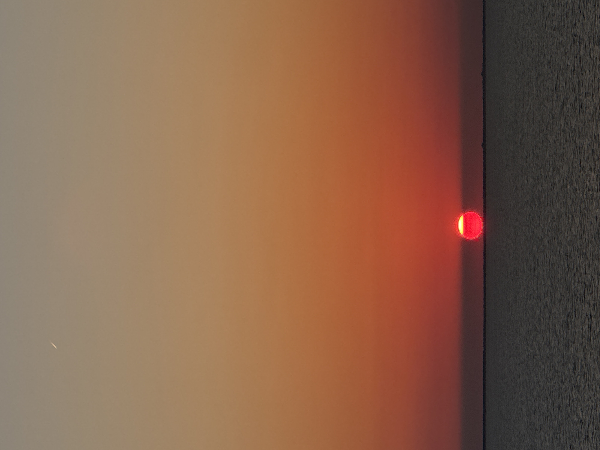 A low-angle, full-frame shot captures a sunset over a body of water. The sun, a vibrant red orb with a bright yellow core, is positioned just above the horizon line, partially submerged. The sky transitions from a pale peach at the top to a deep fiery orange and red as it approaches the sun. A faint, white contrail from an airplane is visible in the upper left quadrant of the sky. The water in the foreground is a dark, textured expanse. The horizon line appears straight and flat, with a subtle, indistinct landmass visible in the far distance. The scene evokes a sense of calm and natural beauty.