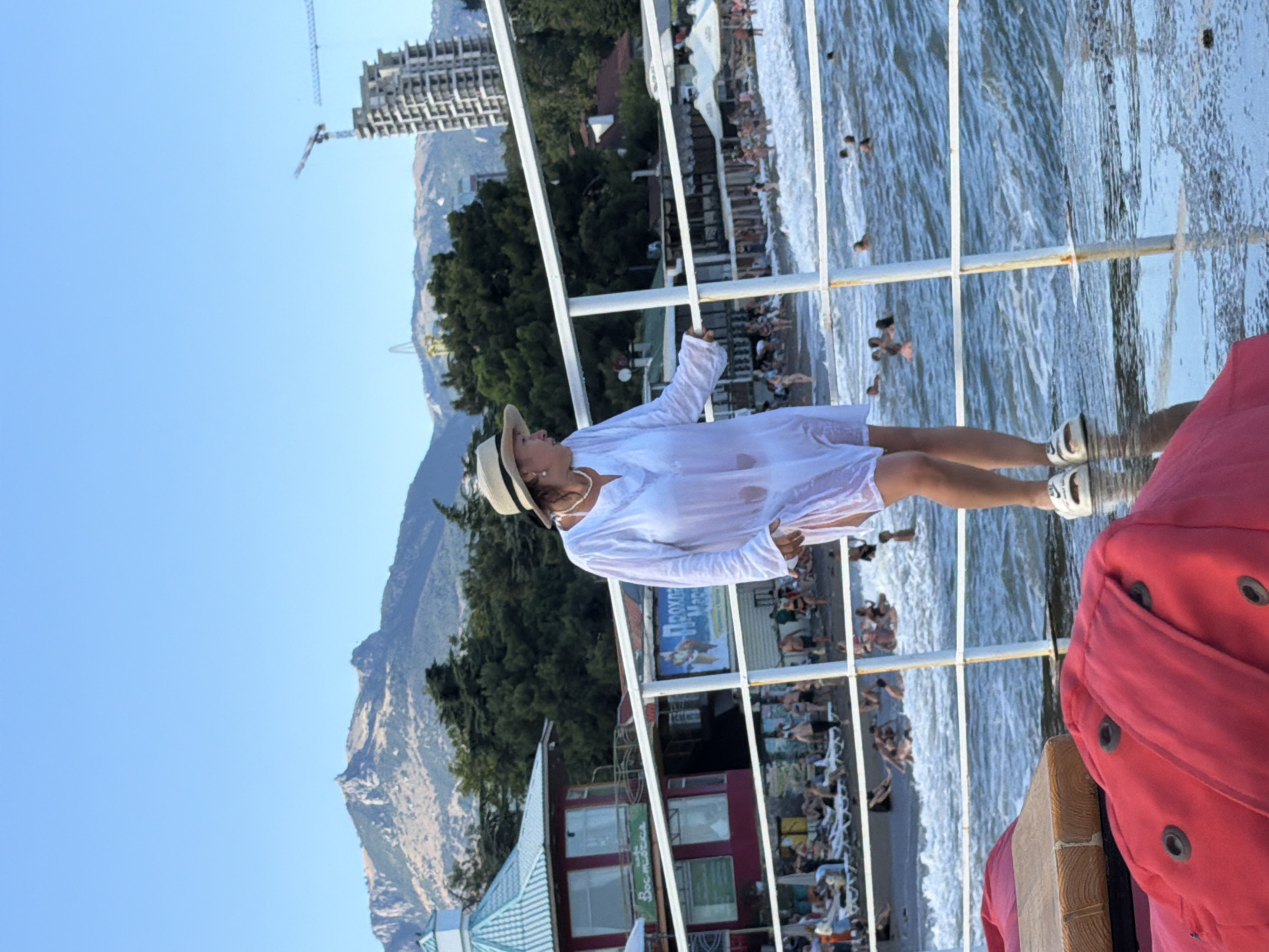 A woman stands on a pier in Alushta, Ukraine, wearing a white cover-up and a hat. The background shows a beach with people swimming and sunbathing, mountains, and buildings. The sky is clear and blue, and the water is choppy.
