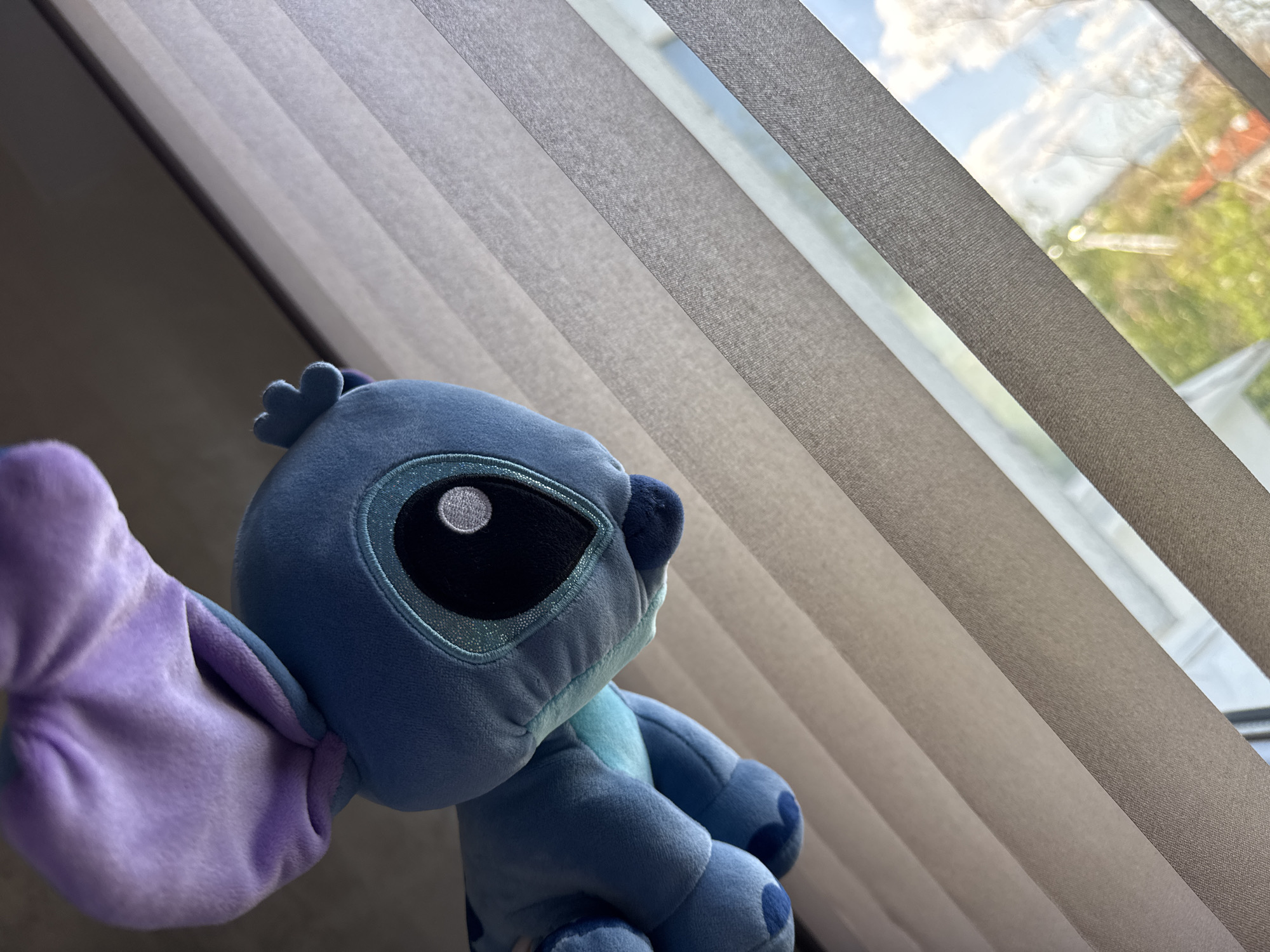 The image shows a Stitch plush toy sitting in front of a window with closed vertical blinds. Outside, through the gaps in the blinds, one can see a blurry view of trees and a sky with clouds. The location appears to be in a residential setting in the city of Unknown, Unknown.
