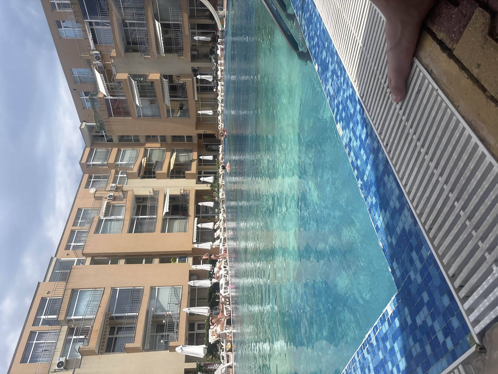 This image depicts a view of a resort-style swimming pool and apartment complex, likely located in Nesebar, Bulgaria. The complex features multiple floors of apartments with balconies, beige-colored facades, and numerous windows. White umbrellas and lounge chairs are arranged around the perimeter of the pool, with several people relaxing on the loungers. Others are present in the bright blue swimming pool. The pool area is bordered by blue tiled edging and a white slatted drain grate. A person's foot is visible in the lower right corner of the image, resting on the grate. The sky above is partly cloudy.