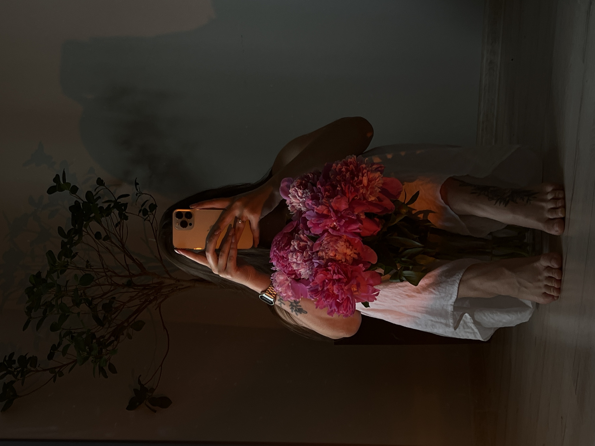 A person is taking a selfie in a room. The person is seated on the floor, wearing a white dress. They are holding a smartphone in their right hand, with their left hand also visible, partially obscured by the phone. A large bouquet of pink peonies is placed in front of them, in a glass vase. A potted plant with green leaves is visible behind the person. The room has a light-colored wall and a wooden floor. Shadows are cast on the wall, including a silhouette of the person and the plant. There is a watch on the person's left wrist. The lighting suggests it is either late afternoon or early evening, with warm light illuminating the scene from the side. The overall impression is one of quiet contemplation. The image was captured in Ternopil, Ukraine.