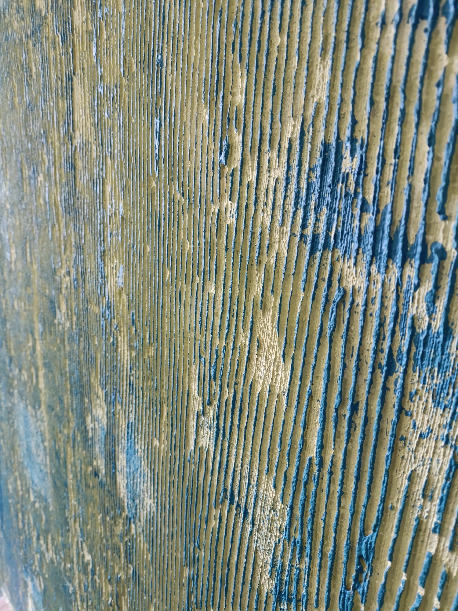 The image displays a close-up, abstract view of a textured surface. The surface is characterized by a series of parallel, vertical ridges and grooves. The dominant colors are a muted, yellowish-green and a deep blue. The texture appears rough and uneven, with areas where the blue paint seems to have chipped or worn away, revealing the yellowish-green beneath, or vice versa. The lighting suggests the texture is emphasized, with shadows falling into the grooves. The perspective is angled, giving a sense of depth and texture. The image is a detail shot, focusing on the pattern and material of the surface, with no discernible context of a specific location within a city.