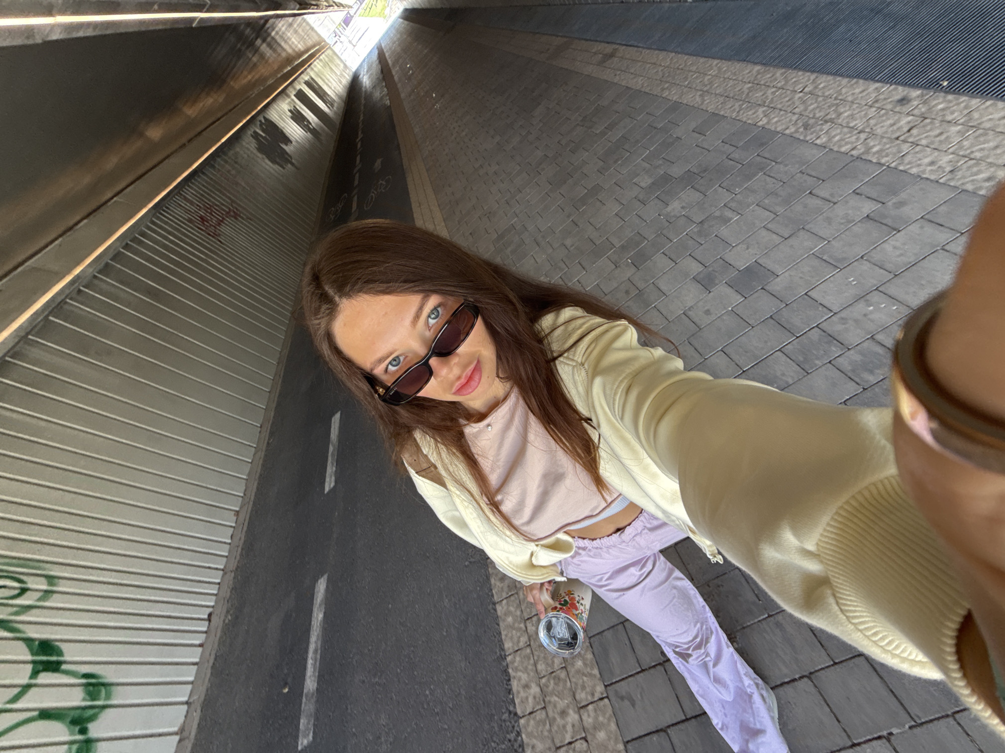 A young woman takes a selfie in an underpass in Tel Aviv, Israel. She wears sunglasses, a light pink top, a yellow jacket, and lavender pants. She holds a reusable cup in her left hand. The underpass has a metal corrugated wall on one side and a gray brick walkway on the other. The walkway has a bike lane marked on it. The background shows the entrance to the underpass, suggesting an exit to the street level.
