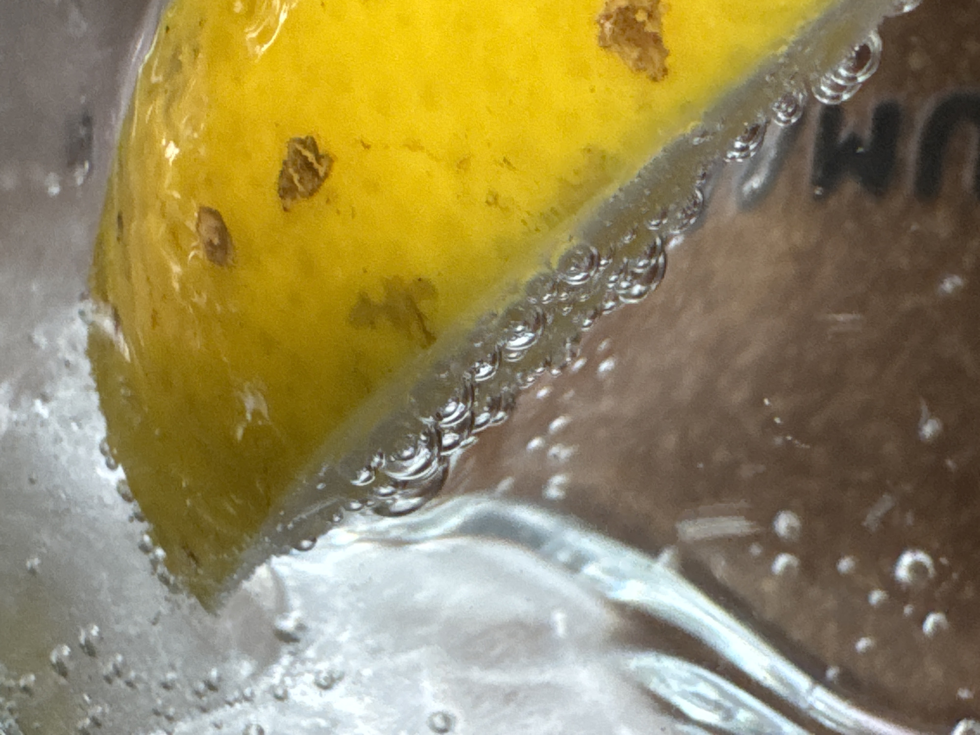 A close-up shot shows a slice of yellow lemon submerged in a clear, bubbly liquid, likely soda or sparkling water. Small air bubbles cling to the lemon's surface along the cut edge. The lemon has some blemishes or spots on its skin. The background is blurry, but part of a word ending in "UM" is visible. The location in the city of North Kuta, Indonesia, cannot be determined based solely on this image.