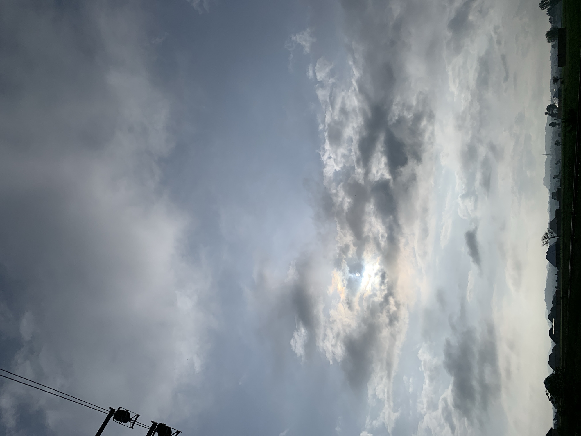 The image shows a view of the sky over Kuru Karama, Nigeria. The sky is filled with a mix of dark and light clouds. The sun is partially obscured by the clouds, creating a diffused light. In the foreground, silhouetted against the sky, are several structures that appear to be houses with thatched roofs, as well as some trees. The landscape appears to be rural, with hills visible in the distance. The overall impression is of a slightly overcast day, possibly late afternoon or early morning. A utility pole with wires is visible on the left side of the frame, indicating the presence of infrastructure in the area.