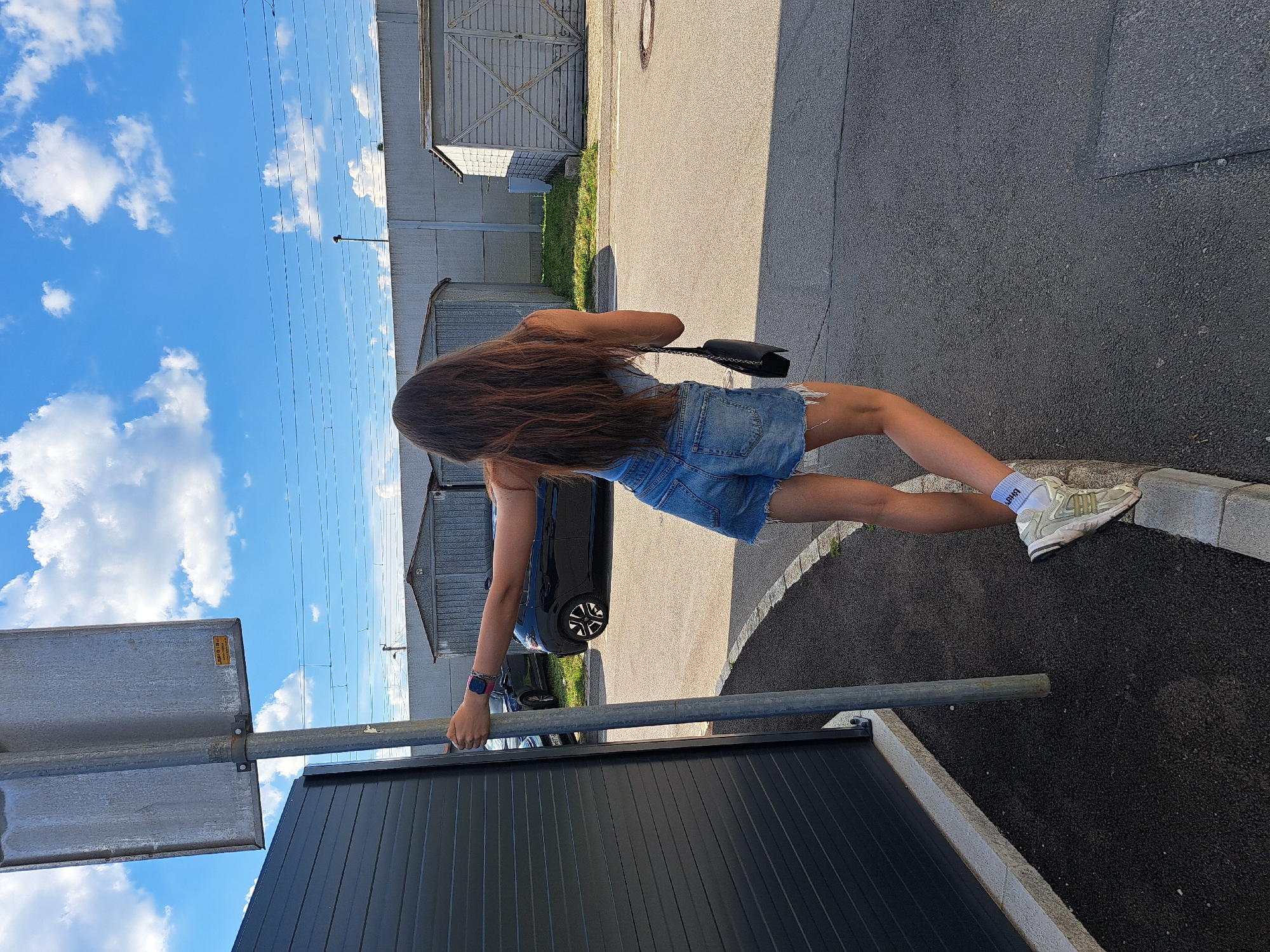 A woman with long brown hair, wearing a blue tank top and denim shorts, stands outdoors next to a metal pole, holding onto it with her left hand. She is facing away from the camera, looking towards the right. She has a black purse slung over her right shoulder and is wearing white socks and light-colored sneakers. Her right foot is propped up on a curb. In the background, there are two black cars parked on an asphalt surface, several grey metal sheds, a concrete wall, and a blue sky with white clouds. Power lines are visible in the sky. The scene appears to be in an urban or industrial area, possibly a parking lot or an alleyway.