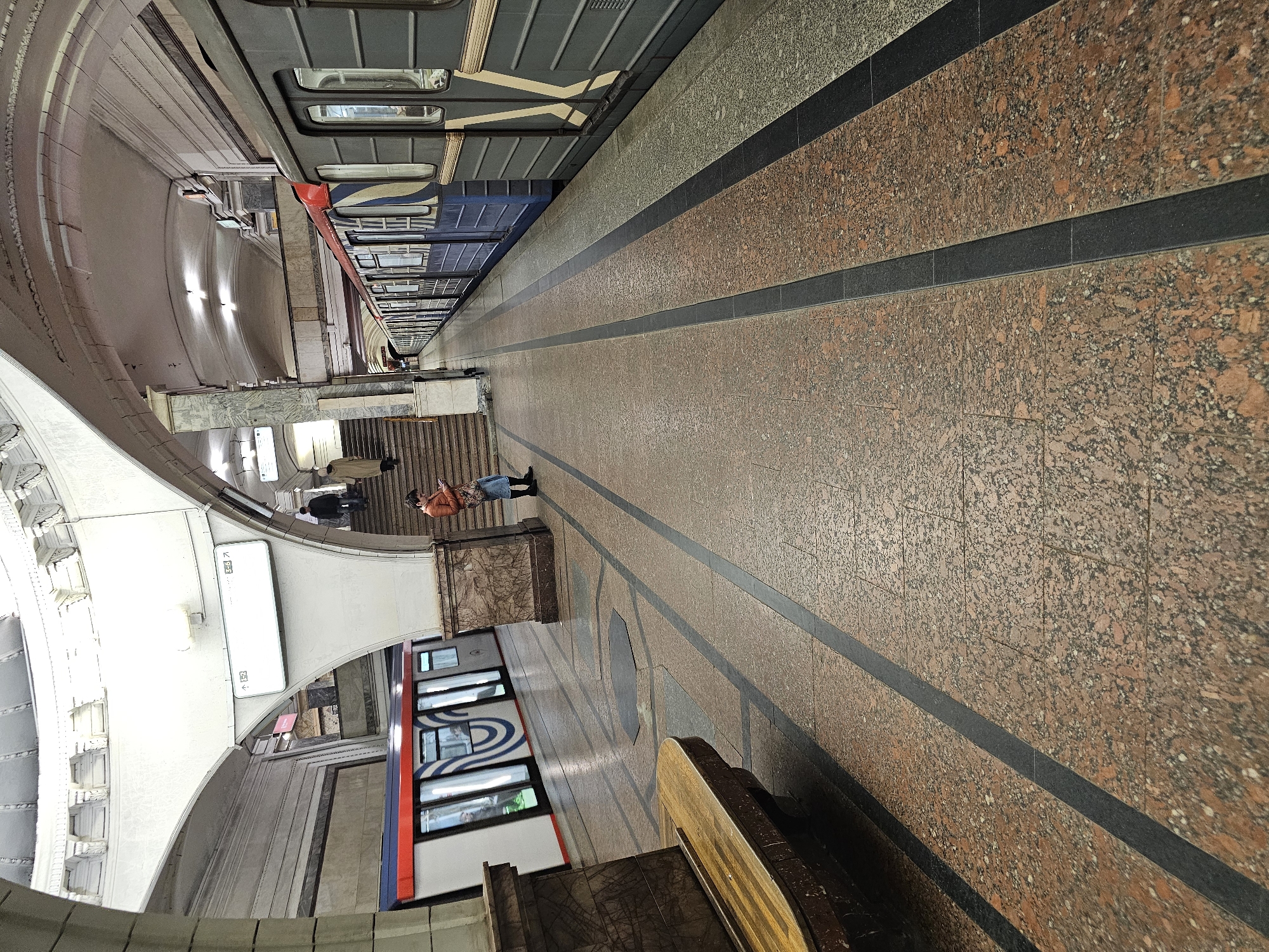 The image depicts a subway station platform in Moscow, Russia, characterized by its polished granite flooring in shades of brown and black. Two trains flank opposite sides of the platform. The one on the left exhibits red and blue exterior design elements. The platform includes seating, station signage, and stairs to an upper level. Several people are on the platform: one appears to be observing a train, and others are moving toward the staircase. The architecture includes arched ceilings and detailed architectural moldings.