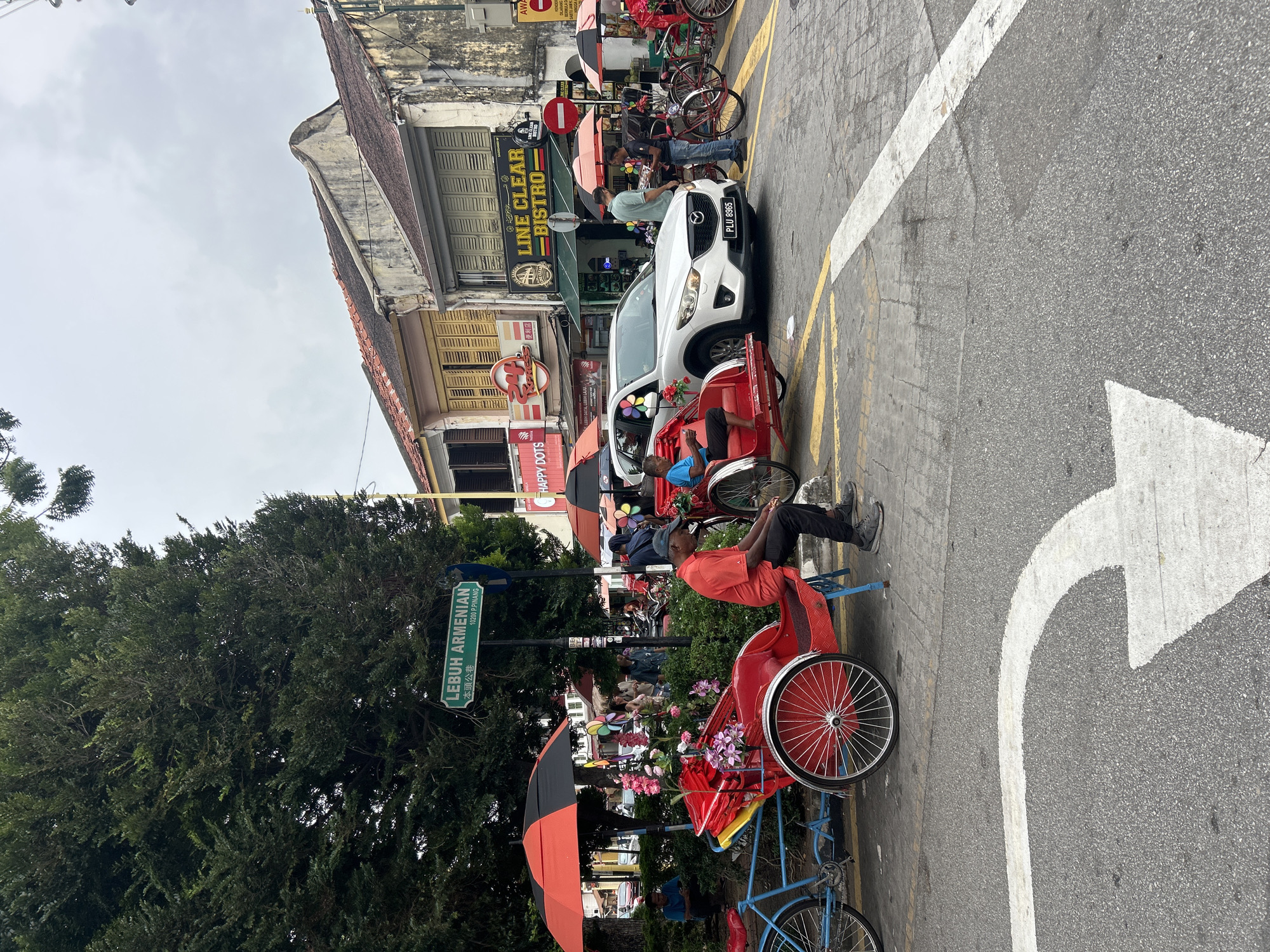 This image captures a street scene in George Town, Malaysia, specifically on Lebuh Armenian. In the foreground, a red and blue pedicab is parked on the side of the road. A man in an orange shirt and dark pants sits on the pedicab's seat, looking down. The pedicab is decorated with pink and purple flowers. Adjacent to it, another pedicab, this one primarily red, is visible, occupied by a person in a blue shirt.
Behind the pedicabs, a white SUV with license plate PLU 8965 is parked in the street. Further down the road, several people are milling about, and various shops line the street, including "LINE CLEAR BISTRO." The architecture features multi-story buildings with traditional shutters and weathered facades. A street sign clearly reads "LEBUH ARMENIAN" and "10200 P.PINANG." Overhead, a large tree with dense green foliage partially obscures the sky, which appears overcast. The ground is paved with asphalt and marked with yellow and white traffic lines.