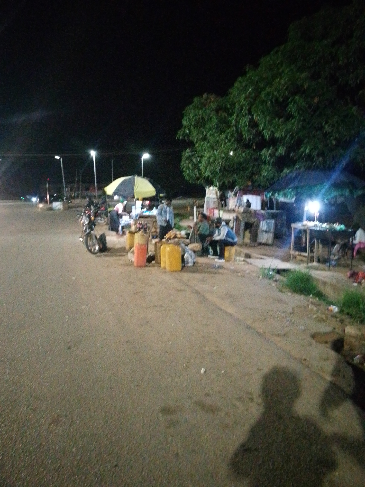 An outdoor scene at night depicts a roadside market with several people and merchandise. The foreground is an asphalt road with the shadow of the photographer visible. To the right of the road, a dirt area contains makeshift stalls and a large tree overhead. Several individuals are present, some seated and others standing near the stalls. Merchandise includes large yellow containers, possibly for liquids, and other items on display tables. A motorcycle is parked to the left of the market. Streetlights illuminate the area, casting a dim glow on the scene.