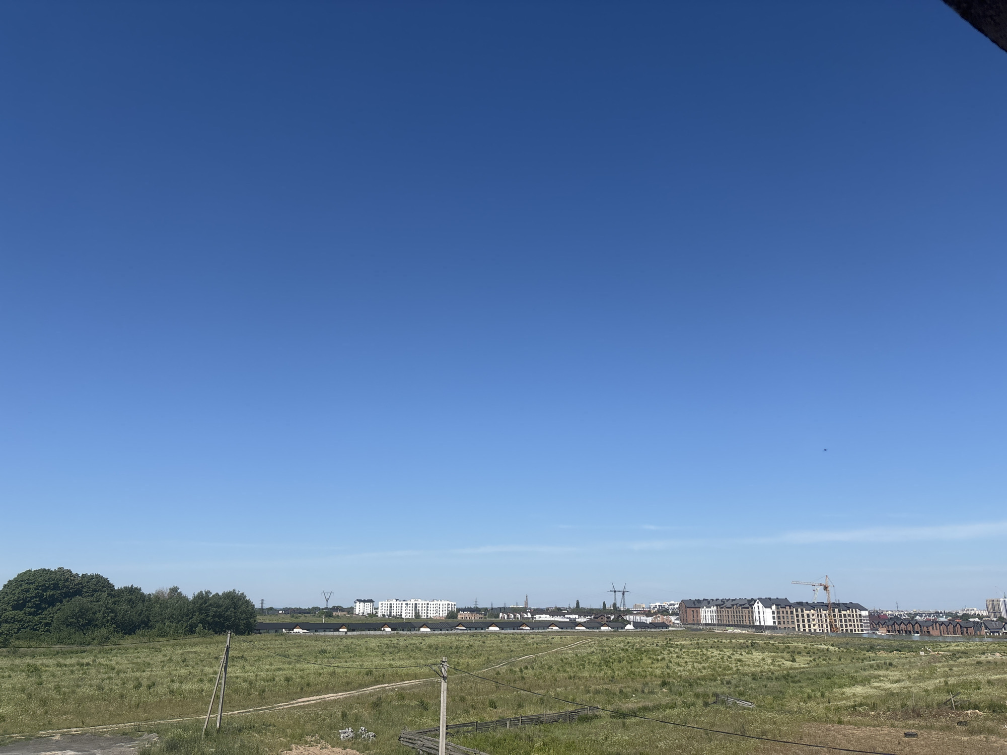 The image shows a wide, panoramic view of a semi-rural landscape under a clear blue sky. In the foreground, a grassy field with scattered wildflowers and some patches of dry grass stretches towards the horizon. A wooden pole with electrical wires crosses the field diagonally from the lower left to the upper right. A dirt path can be seen winding through the field.
In the middle ground, a line of buildings representing a developing residential area stretches across the scene. These buildings appear to be modern townhouses and apartment complexes with dark roofs. To the far left, a dense cluster of green trees provides a stark contrast to the open field. Further into the background, the skyline features more buildings, power lines, and a construction crane, suggesting ongoing development.
The location is identified as Strumivka, Ukraine. The scene depicts a transitional area between undeveloped land and urban expansion, characterized by a clear, bright day. There are no people visible in the image.