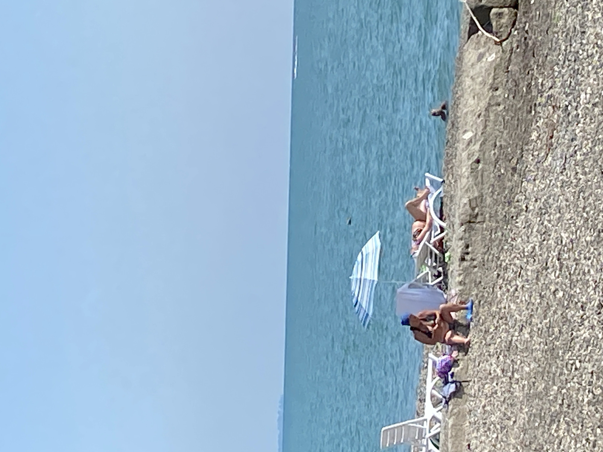 The image depicts a coastal scene in Chabkwara, Abkhazia. The foreground is a stony beach with several beach chairs and umbrellas. In the middle ground, two individuals are visible. One, wearing a blue headscarf and a bikini, is seated on the beach, looking down at their hands. Another person is lying on a lounge chair under an umbrella, facing away from the camera. Further out in the turquoise water, a person is partially submerged, with only their head and shoulders visible. In the distance, a faint line of land is visible on the horizon under a clear blue sky. A small white boat or jet ski is creating a wake on the far right of the water.