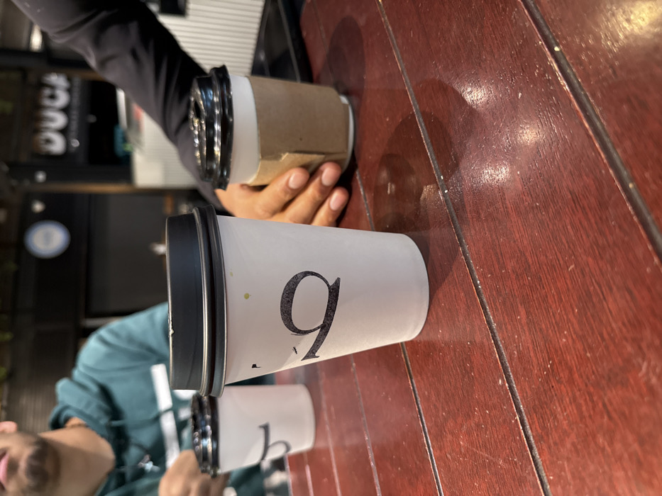 Two coffee cups sit on a red-brown wooden surface. The first cup has a large black "9" on it, with a black lid. The second cup has a similar design but the number is obscured. A hand holds a third cup with a brown sleeve. Part of a person's face is visible in the bottom left, wearing a green and white top. Another person's arm, clad in black, is partially seen at the top left. The background appears to be a coffee shop interior.