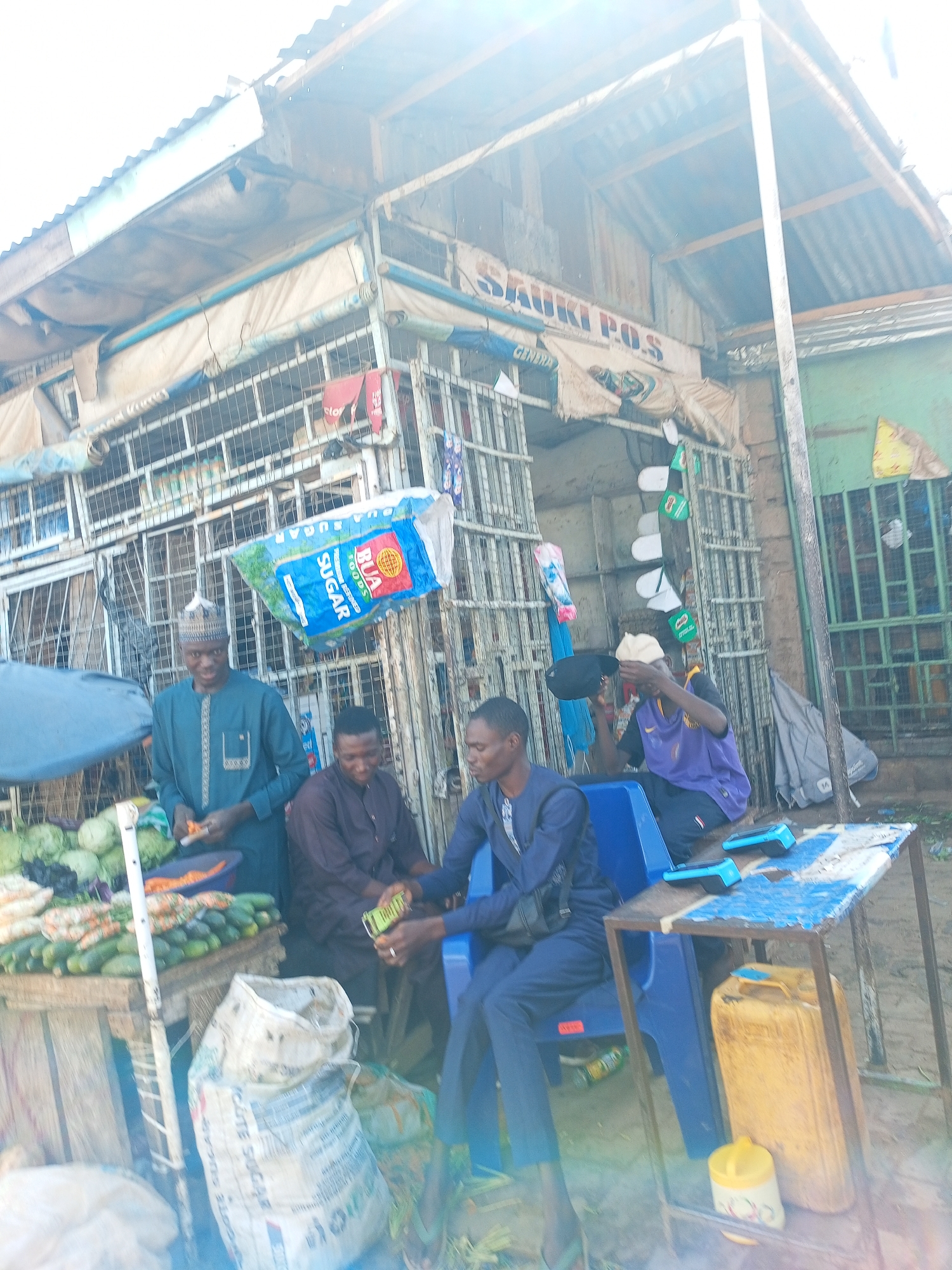 This daytime photograph captures a vibrant market scene, likely a roadside stall, under bright sunlight which causes a strong lens flare. The setting features a makeshift structure with a corrugated metal roof, supported by metal frames and covered partially with tarpaulin.
At least four men are visible. On the left, a man in a teal-colored kaftan and a kufi hat stands behind a wooden stall laden with fresh vegetables, including cabbages, carrots, and cucumbers. He appears to be preparing vegetables. Next to him, on the right, two men are seated on blue plastic chairs. One man wears a brown traditional outfit, and the other a dark blue one. Further to the right, another man in a purple shirt and a light-colored cap is also seated on a blue plastic chair, appearing to be focused on something in his hands or lap.
Prominently displayed, a large blue and white bag labeled "BUA FOODS PREMIUM REFINED SUGAR" hangs from the metal frame. Nearby, a large white sack, which OCR identifies as "GOTE SUGAR", rests on the ground. Various other goods, including packaged items and possibly shoes/sandals, hang from the stall's structure. On a small, makeshift table made of metal and wood, two light blue point-of-sale (P.O.S.) terminals are visible. Underneath the table, a yellow jerry can and a smaller yellow container are placed.
A sign above the stall clearly reads "SAUKI P.O.S", indicating the presence of a point-of-sale service. Another partial text reads "GENERA". The overall atmosphere suggests an informal commercial area, typical of a local market or roadside business.
