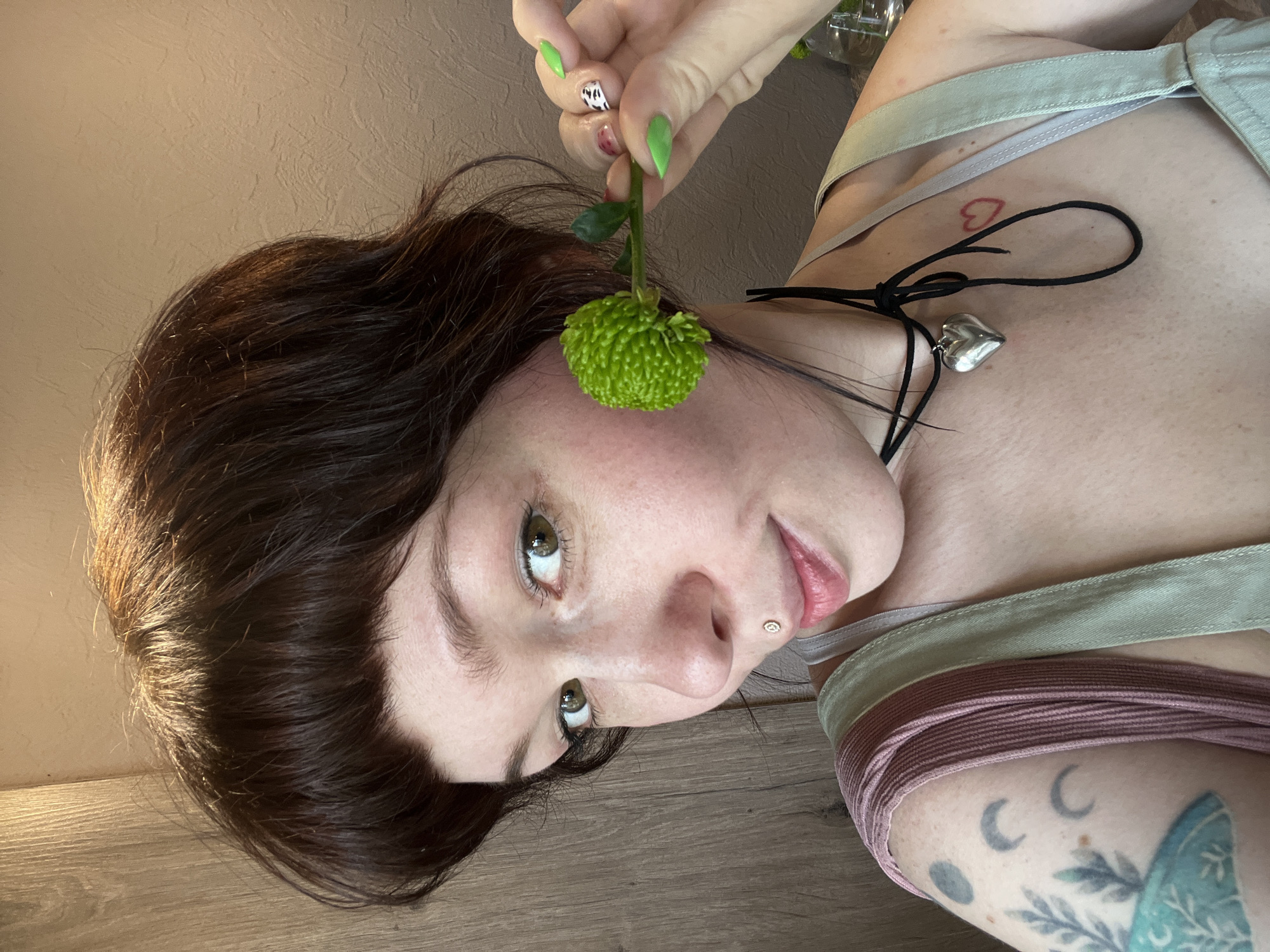 A person is holding a green flower next to their face. They have brown hair with bangs, fair skin, and green eyes. A silver heart-shaped pendant hangs from a black cord around their neck. A small red heart tattoo is visible on their chest. They are wearing a green top and a mauve colored band is visible around their arm. There is a butterfly tattoo on their arm. The setting appears to be indoors, with a beige wall behind the person. The photo is taken from an angle, with the person's head tilted. The location of the city of Chaikovsky, Russia is unknown, and cannot be extracted from the image.
