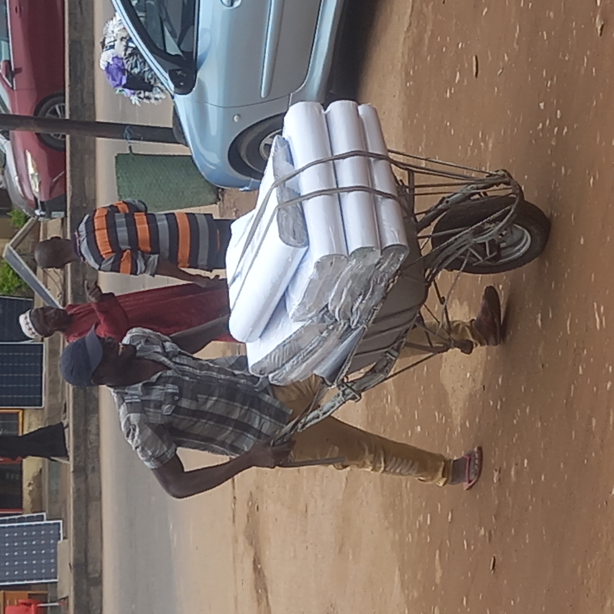The image depicts a street scene in Sardauna, Nigeria. A man in a blue baseball cap, plaid shirt, and khaki pants is pulling a wheelbarrow loaded with tightly packed, white rolls, possibly paper or fabric, secured with ropes. Two other men are nearby. One is wearing a red robe and a white cap, and the other is wearing a horizontal striped shirt. Various objects are in the background, including cars, a green patch of grass, and what appears to be a display of solar panels. The ground is a light brown color, likely dirt or pavement.