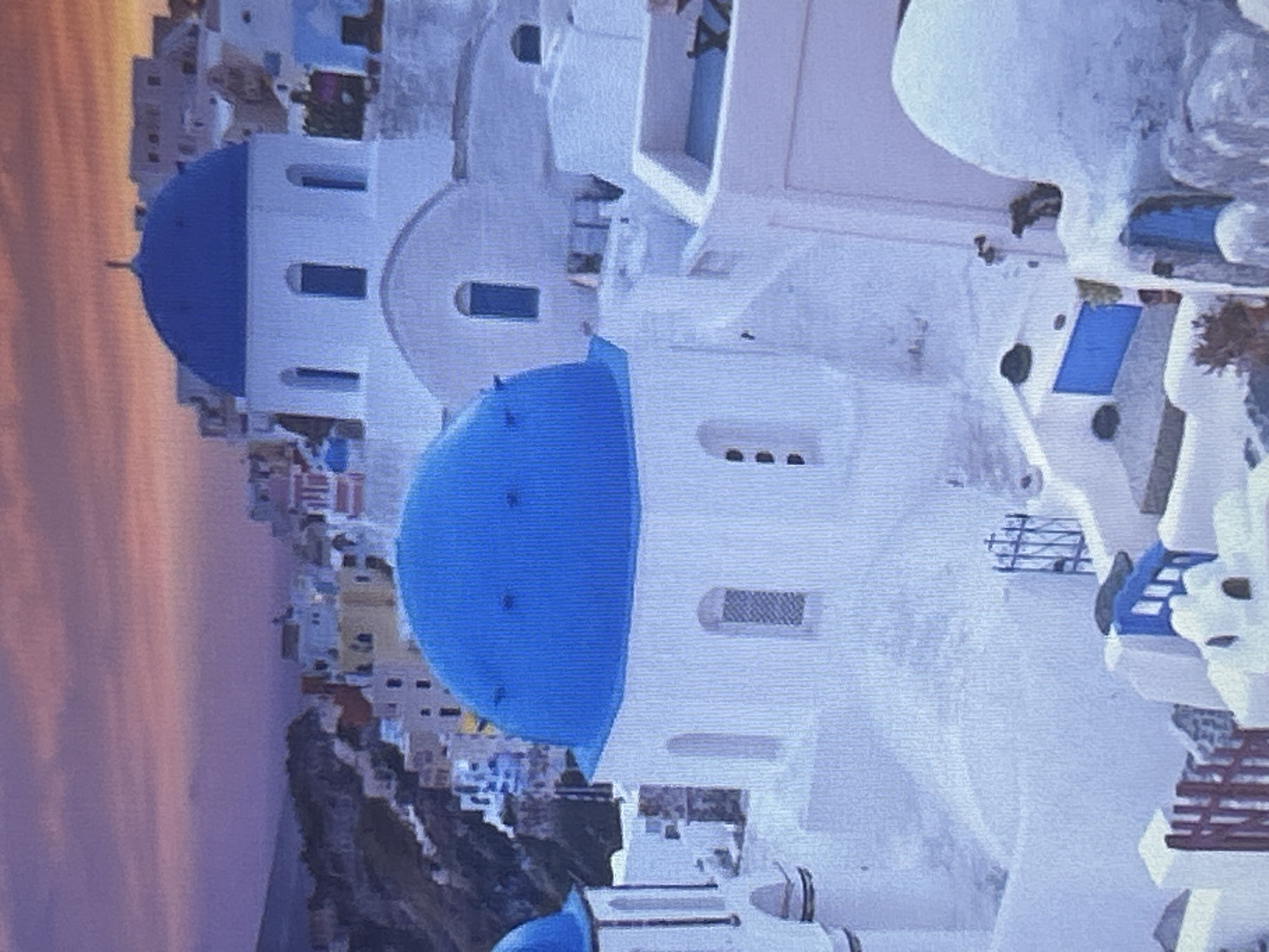 The media file depicts a picturesque scene of a cliffside village with distinctive white buildings and blue-domed structures, characteristic of a Greek island like Santorini. The architecture features cubic houses with flat roofs, whitewashed walls, and blue accents, including window frames and doors. Several blue domes are visible, suggesting churches or chapels. The village is built on a steep incline, overlooking a body of water. In the background, the sky displays soft hues of orange and purple, indicative of either sunrise or sunset. The overall impression is serene and idyllic, evoking a sense of tranquility. There is no visible text in the image, and no people or specific activities are clearly discernible. The scene is bathed in the warm, diffused light of dusk or dawn. The location context provided, Sofia, Bulgaria, appears incongruous with the visual evidence of a Mediterranean island village.