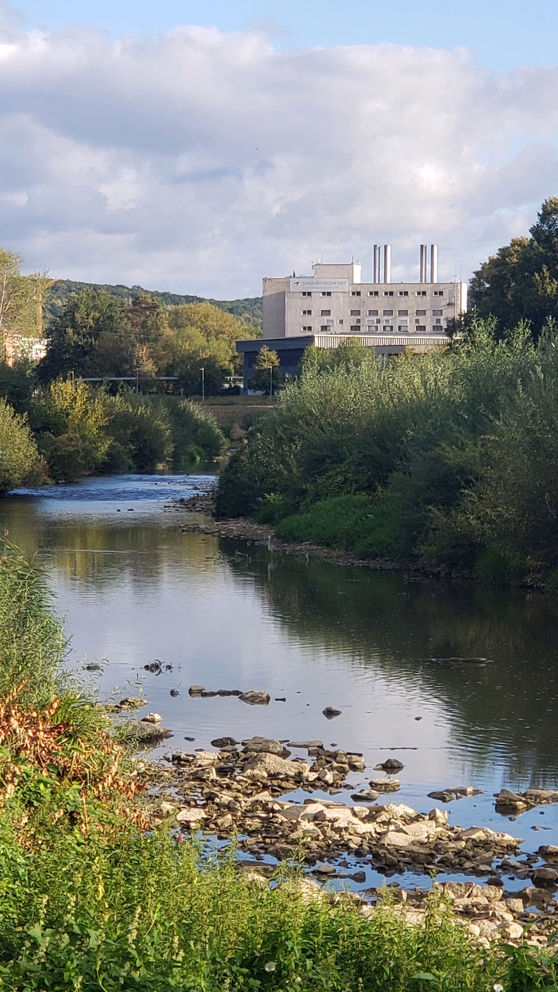 A vertical, eye-level, long shot captures a scenic view of the Torysa River in Prešov, Slovakia. The river, with its shallow, rocky bottom and calm, reflective surface, flows from the foreground into the background. Lush green vegetation lines the riverbanks, with a mix of trees and bushes creating a natural, unmanicured edge. In the background, a modern, multi-story building with a flat roof and several ventilation stacks stands out against a backdrop of rolling hills covered in trees. The sky is partly cloudy, with a mix of sunlight and shadows playing across the landscape. No people are visible in the scene.