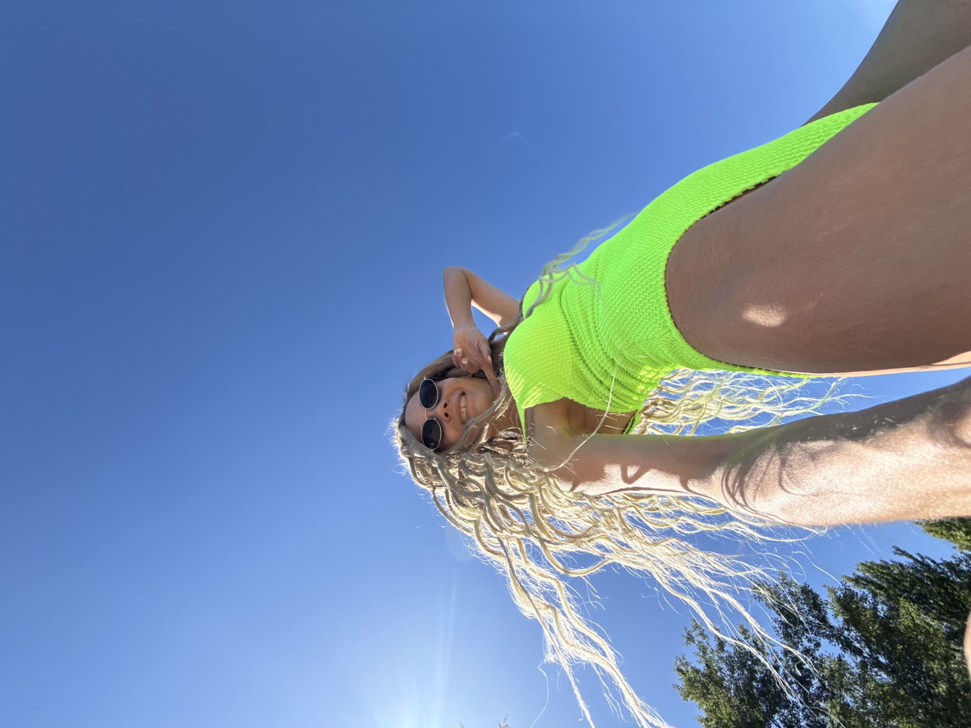 An outdoor, low-angle photograph captures a young woman from below, set against a bright, clear blue sky in Шебунино, Russia. The woman has long, highly textured, light-blonde hair, which is windblown and extends upwards and to the left. She is wearing dark, circular sunglasses and a vibrant neon green swimsuit or leotard, characterized by a distinct waffle-knit texture. Her face is directed towards the camera, and she is smiling broadly, revealing her teeth. Her right arm is raised, with her hand positioned behind her head. Intense sunlight, originating from the upper left, casts a prominent lens flare and sharp shadows on her skin and hair. Dark green tree foliage is visible at the extreme bottom left of the frame.