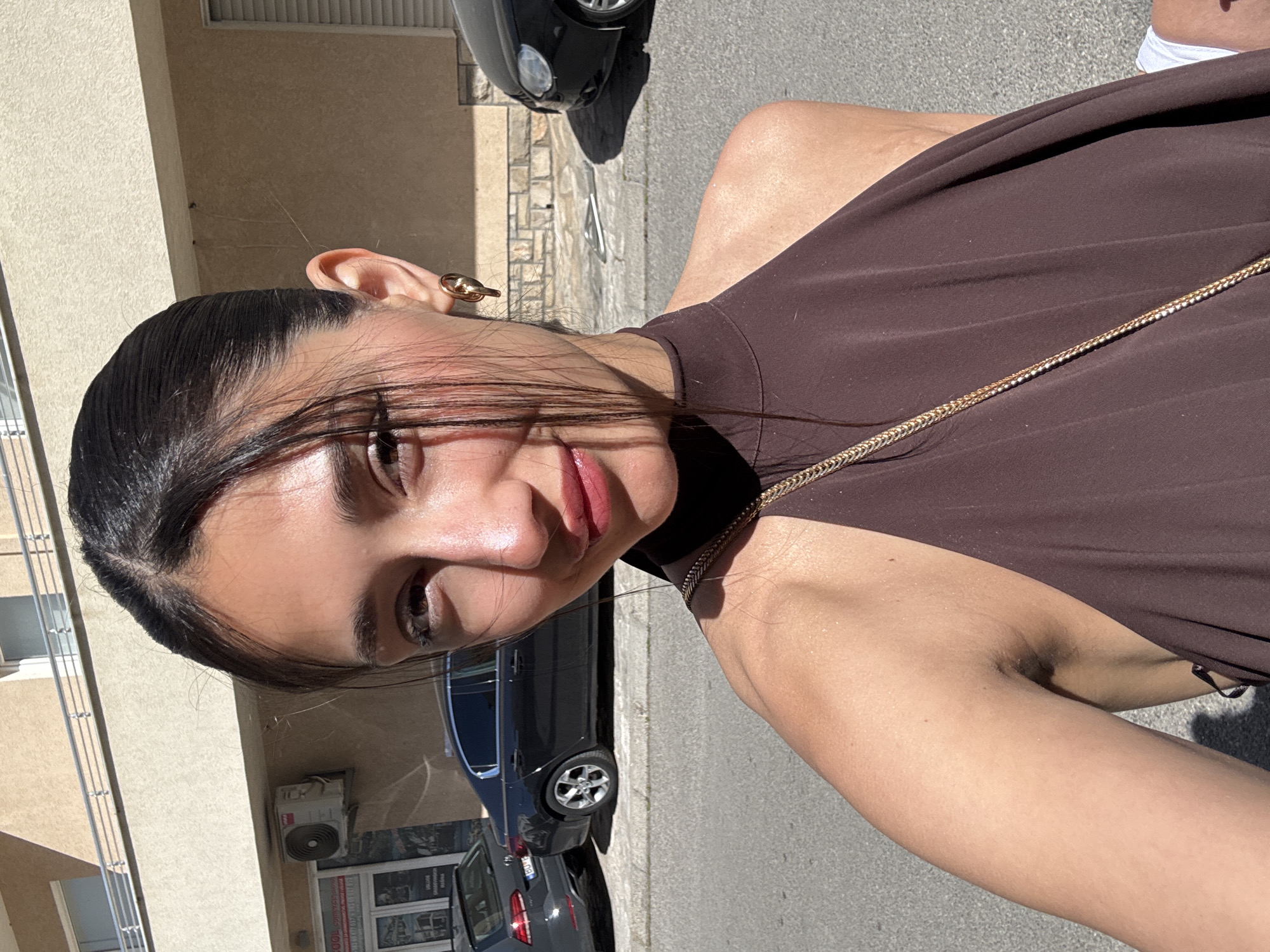 A woman with dark hair and light skin is positioned on a city street in Budva, Montenegro. She is wearing a brown halter top with a gold chain. Her hair is pulled back, and she has an earring in her left ear. Behind her are several parked cars, the side of a building, and a storefront with signage in Cyrillic. The street is paved and appears to be in a residential or commercial area. The lighting suggests it is daytime with sunlight.