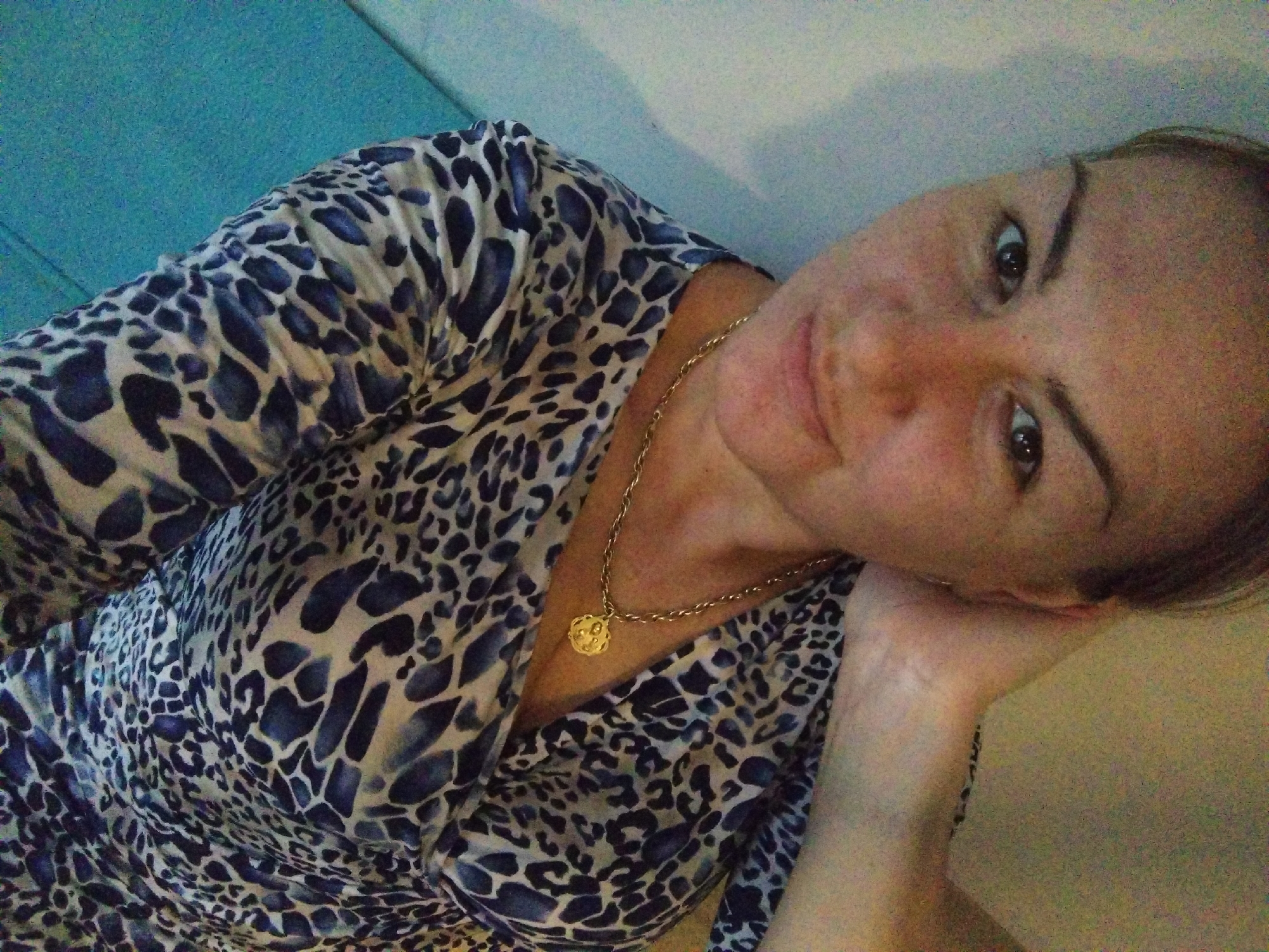 A woman with fair skin and brown hair, wearing a blue and white patterned blouse and a gold necklace with a round pendant, is leaning her head on her hand. The background appears to be a room with a blue wall on the left and a white wall on the right. The location is unknown and there is no indication that the image was taken in the city of Stara Zagora, Bulgaria.