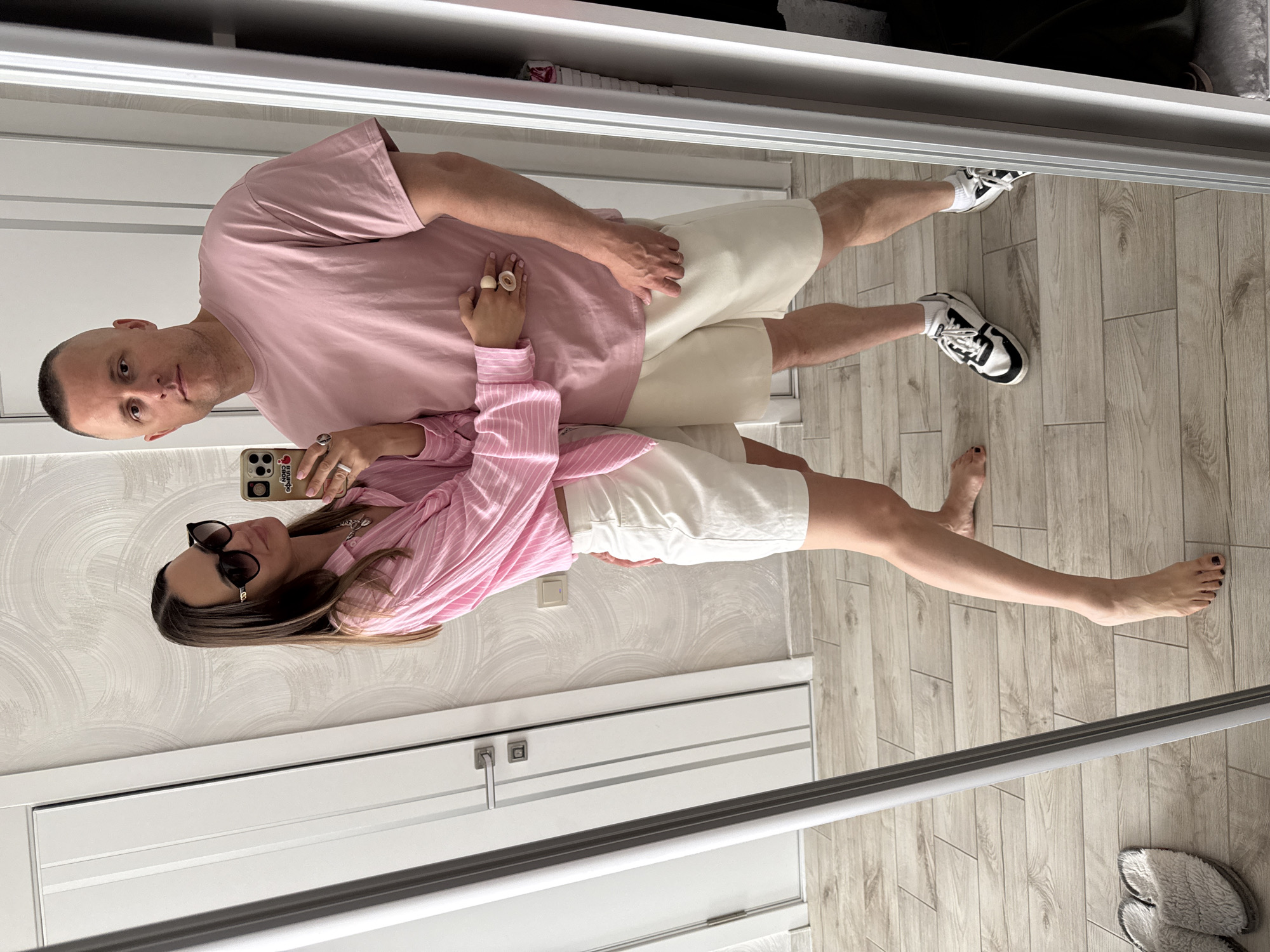 A couple takes a mirror selfie in a well-lit room with light wood flooring. The woman, on the left, wears a pink and white striped collared shirt tied at the waist and white shorts. She has dark hair, sunglasses, and is wearing rings on her fingers. The man, on the right, wears a pink t-shirt and light beige shorts. He has short hair and is wearing rings on his fingers and white athletic shoes with black accents. The room appears to be a home, with a white door and light textured wallpaper visible. The mirror has a silver frame.
