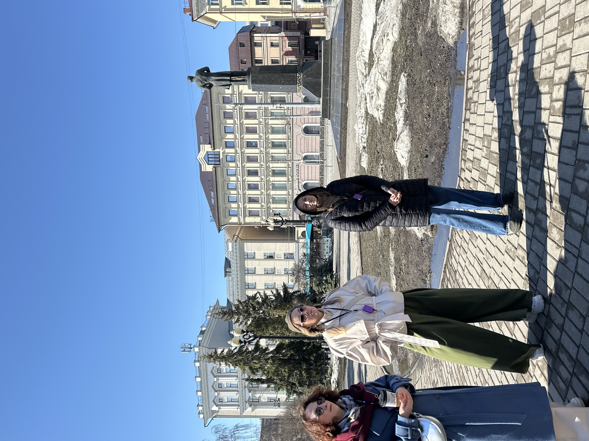 The image shows three people standing on a paved area in front of a statue in Kazan, Russia. The background includes buildings and a clear blue sky. The person in the front is wearing a blue coat and sunglasses, and holding a silver container. The person in the middle is wearing a light-colored coat, dark green pants, and sunglasses. The person in the back is wearing a black jacket, blue jeans and has a hood up. There's a statue of a figure in the background behind them, near the building with multiple windows. The pavement is visible in the foreground, with patches of snow or ice present in the background. Shadows from the people are cast on the pavement.