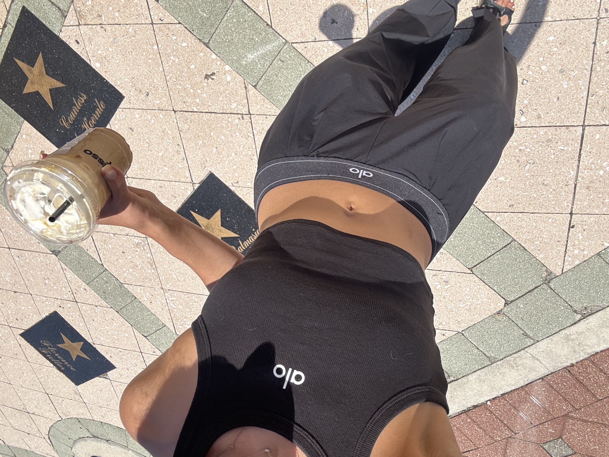 The image shows a person standing on a sidewalk with inlaid stars. The person is wearing a black crop top with the "alo" logo and black pants with the "alo" logo on the waistband. They are holding a clear plastic cup with a beverage and whipped cream. The stars on the sidewalk appear to be part of a walk of fame. One star is inscribed with "Florence Guiller" and another with "Countess Hoernle". The background consists of a patterned sidewalk with pink, green, and tan tiles, as well as shadows from sunlight. The image appears to be taken in a city, likely a tourist area due to the walk of fame. The specific location in Boca Raton, United States of America, is not discernible from the visual information provided in the image.