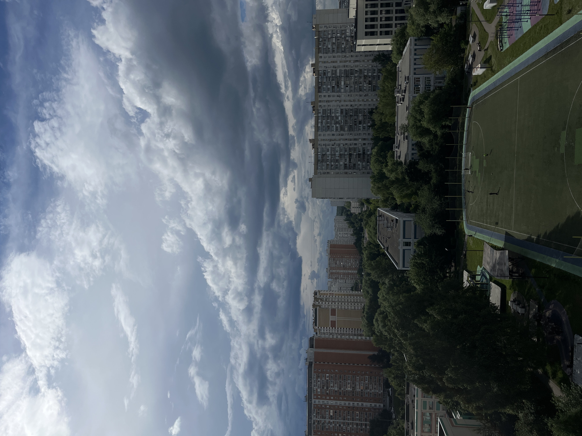 The image captures a view of a residential area in Moscow, Russia, likely from an elevated vantage point such as a building or balcony. Dominating the sky are dramatic, dark storm clouds interspersed with patches of brighter sky, suggesting an impending or recently passed rain shower. Below the sky, a cluster of multi-story apartment buildings forms the urban landscape. The buildings exhibit a typical Soviet-era architectural style, with many floors and numerous windows. Some buildings are taller and more prominent, while others are shorter and set further back.
In the foreground and mid-ground, a significant amount of greenery is visible, primarily dense trees, indicating the presence of parks or courtyards within the urban setting. Among the trees and buildings, a green sports field, possibly a soccer or multi-purpose court, is clearly visible. Several small figures, presumably people, are scattered on the field, some appearing to be engaged in sports. Adjacent to the field, there are playground-like structures with colorful equipment, suggesting an area for recreation. The overall scene portrays a typical urban environment in Moscow, characterized by a blend of residential buildings, abundant greenery, and recreational facilities, under a dynamic and cloudy sky.