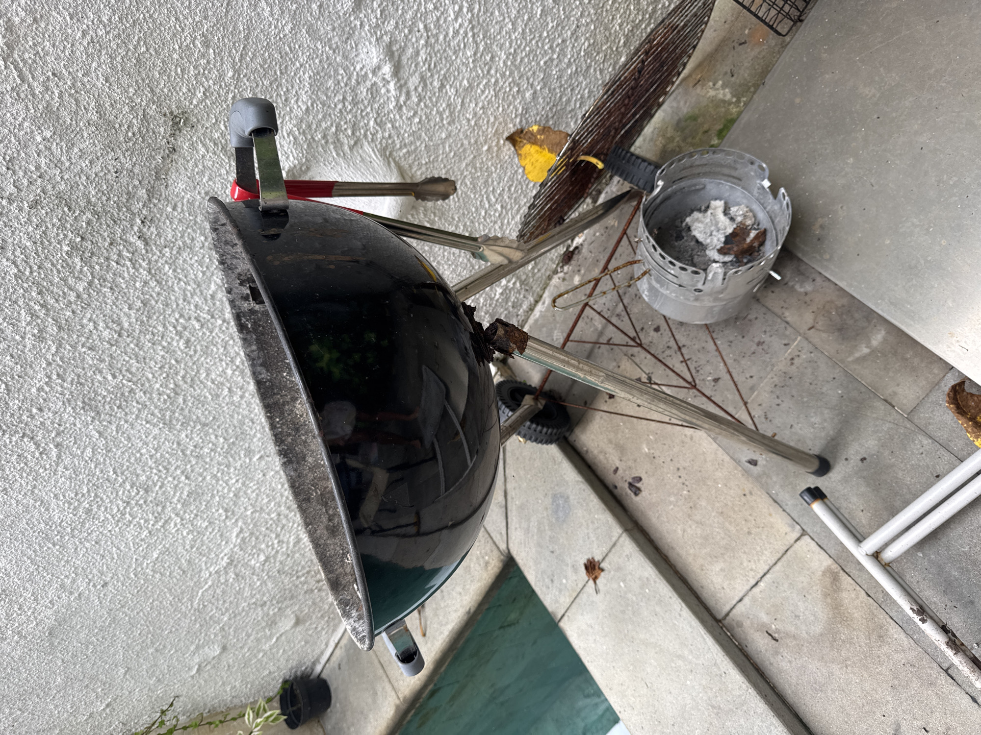The image shows a charcoal grill, with the lid closed, positioned next to a textured wall and a tiled patio or walkway. A chimney starter containing ash is next to the grill. There are grilling tools and a grilling grate nearby. The location appears to be outdoors. The location in the city of Kuta Selatan, Indonesia cannot be determined.