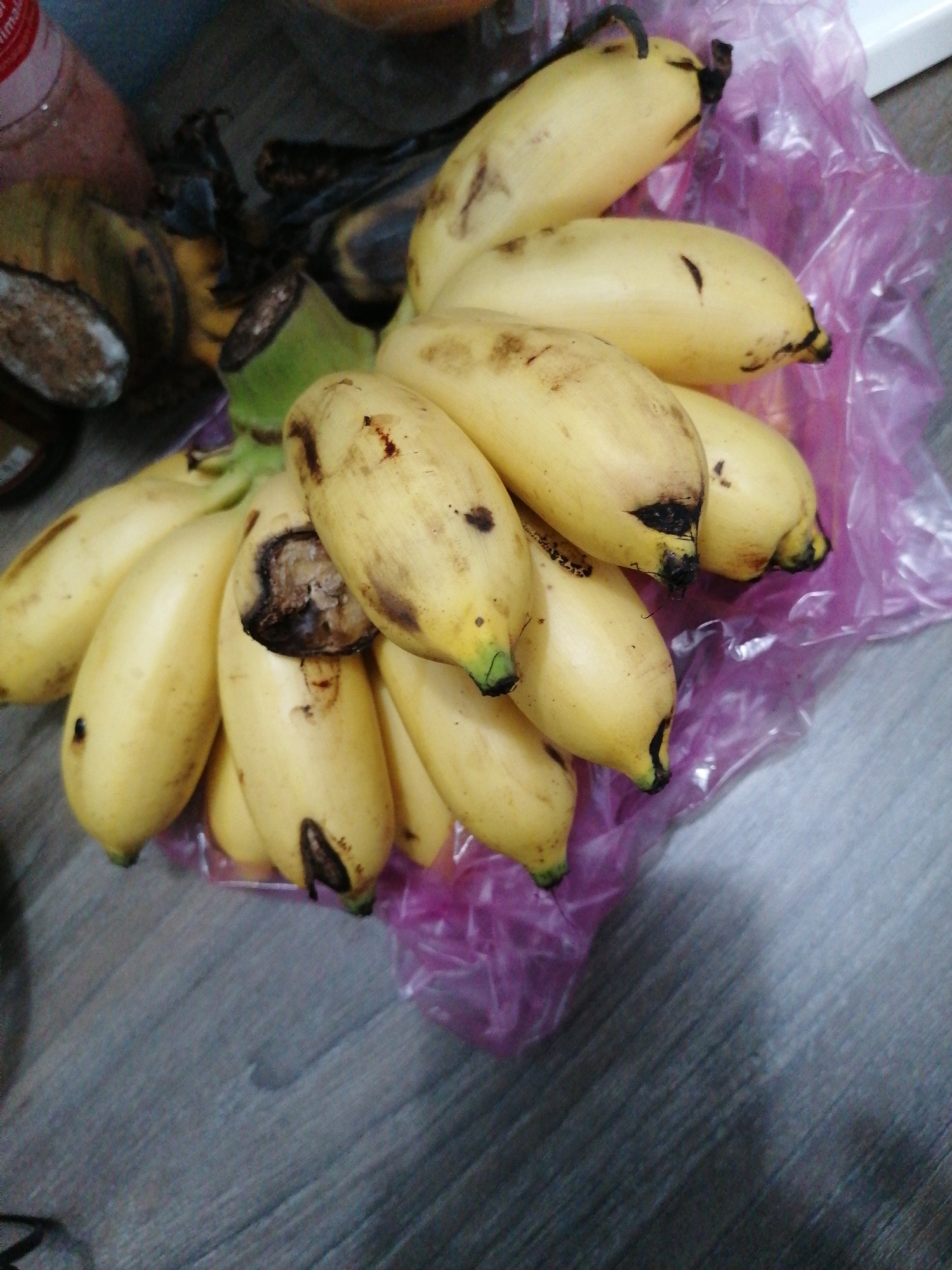 The image displays a bunch of ripe, yellow bananas arranged on a greyish, textured surface, likely a wooden or laminate floor. The bananas are the central focus, mostly bright yellow with various brown spots and marks indicating ripeness, and some showing minor bruises or damage. One banana, prominent in the center, has a large, dark, exposed area where the peel is broken, revealing the discolored fruit beneath.
The bananas are partially resting on and surrounded by a translucent purple plastic bag, suggesting they were recently purchased or are being stored. In the upper left portion of the image, out of focus, are dark, possibly dried banana peels or remnants of a banana stem, along with a partial view of a clear plastic bottle topped with a red cap. No specific activity or event is depicted, and no human presence or discernible emotions are shown. There is no visible text in the image. The provided context indicates the location is Bai Chay Ward, Vietnam.