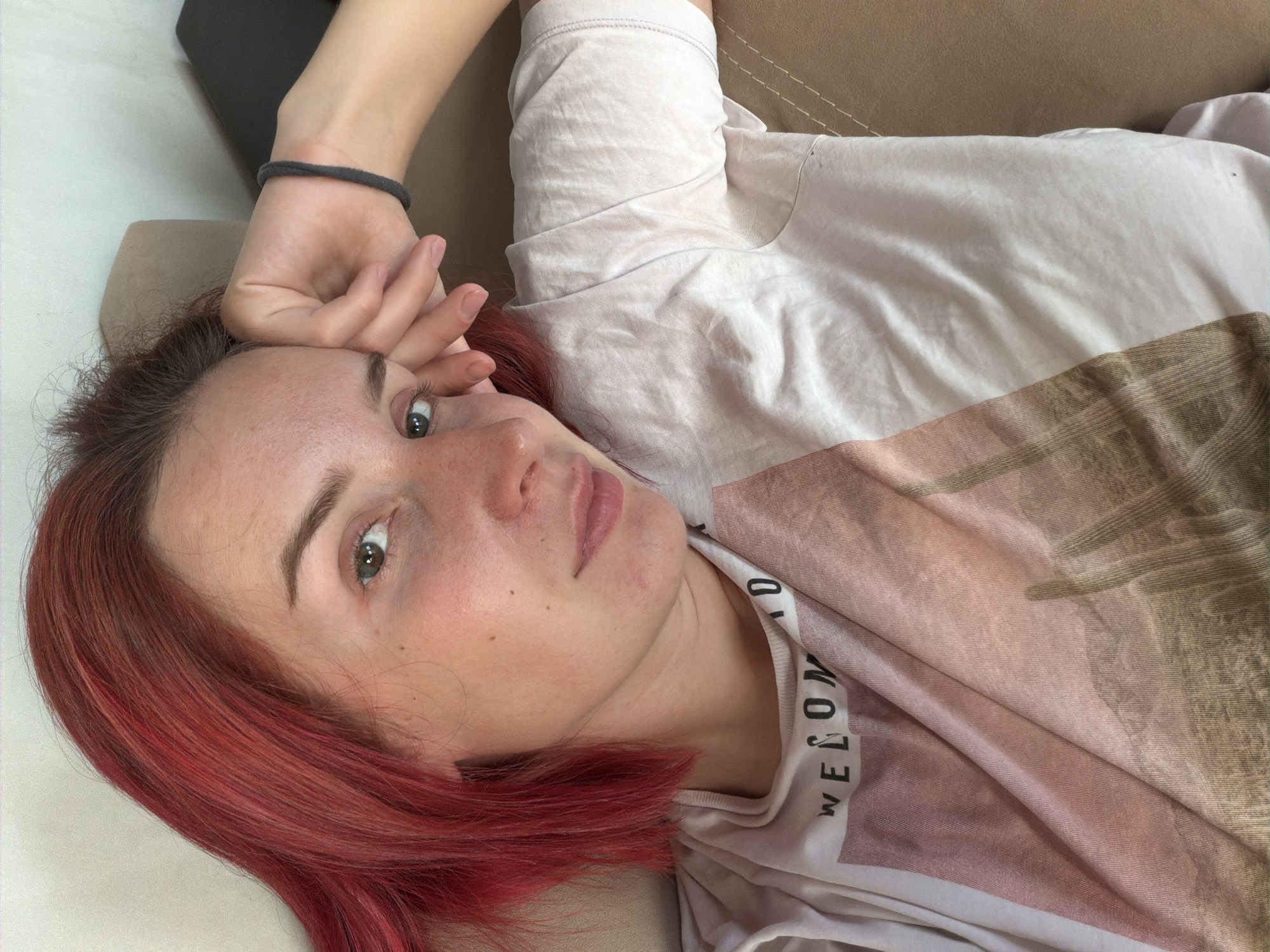 A young woman with red hair is lying on a tan-colored upholstered surface, possibly a sofa or bed. She is facing the camera with a relaxed expression. Her left hand is raised near her forehead, adorned with a black elastic wristband. She is wearing a light-colored t-shirt with a subtle pattern or design. The background is blurred, suggesting a domestic interior setting. The scene is likely taken in Chaikovsky, Russia.