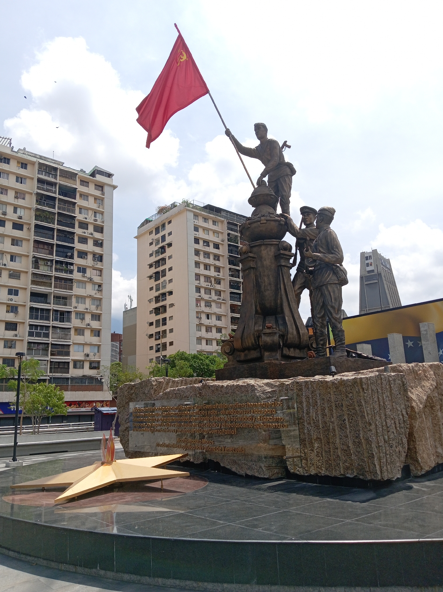 The image depicts a monument in the city of Caracas, Venezuela. The monument consists of bronze statues of three soldiers, one of whom is holding a red flag with a symbol resembling a hammer and sickle. The statues are mounted on a rough-hewn stone base. In front of the monument is a golden star with a flame in the center, set on a dark, reflective circular base. Behind the monument, there are several tall buildings and a partial view of a large yellow, blue, and red flag, possibly the Venezuelan flag. The sky is partly cloudy. The base of the monument appears to have an inscription in Spanish attributed to Nicolas Maduro Moros.