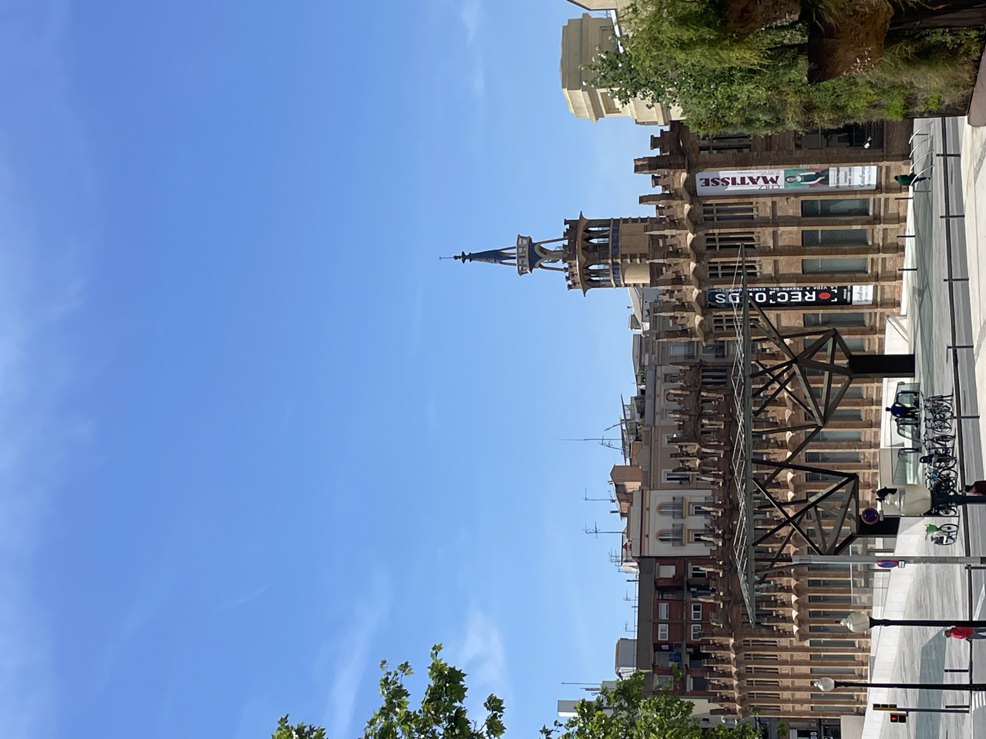 This media file captures a vibrant, sunlit street scene in Barcelona, Spain, on a clear day. The dominant feature is a tall, ornate building with distinctive Modernista architecture, crowned by an elegant spire or tower. This building is characterized by its textured, light brown facade, decorative elements, and numerous windows. The spire itself is slender, dark-colored with lighter accents, and tapers to a point.
To the right of this central building, other urban structures are visible, featuring more traditional architectural styles, some with balconies and rooftop antennas. Green foliage from trees lines the street, adding natural elements to the urban landscape.
The foreground shows a clean, paved street or sidewalk area with parked bicycles, streetlights, and a traffic signal. A large, dark-colored, crisscrossing metal structure spans across the lower part of the image, possibly part of an overhead walkway or support system.
The sky is a bright, clear blue with very few, almost imperceptible, wisps of clouds, indicating excellent weather. The strong sunlight suggests it is daytime, likely late morning or early afternoon.
Visible text includes a large banner on the ornate building displaying "MATISSE" next to an image of a person, indicating an art exhibition or themed establishment. Below this, another banner partially reads "REC.O.DS," likely referring to "RECORDS." Several people are present, though not prominently featured; one is near the parked bicycles, and another is visible further down the street. The overall impression is that of a bustling yet pleasant urban environment.