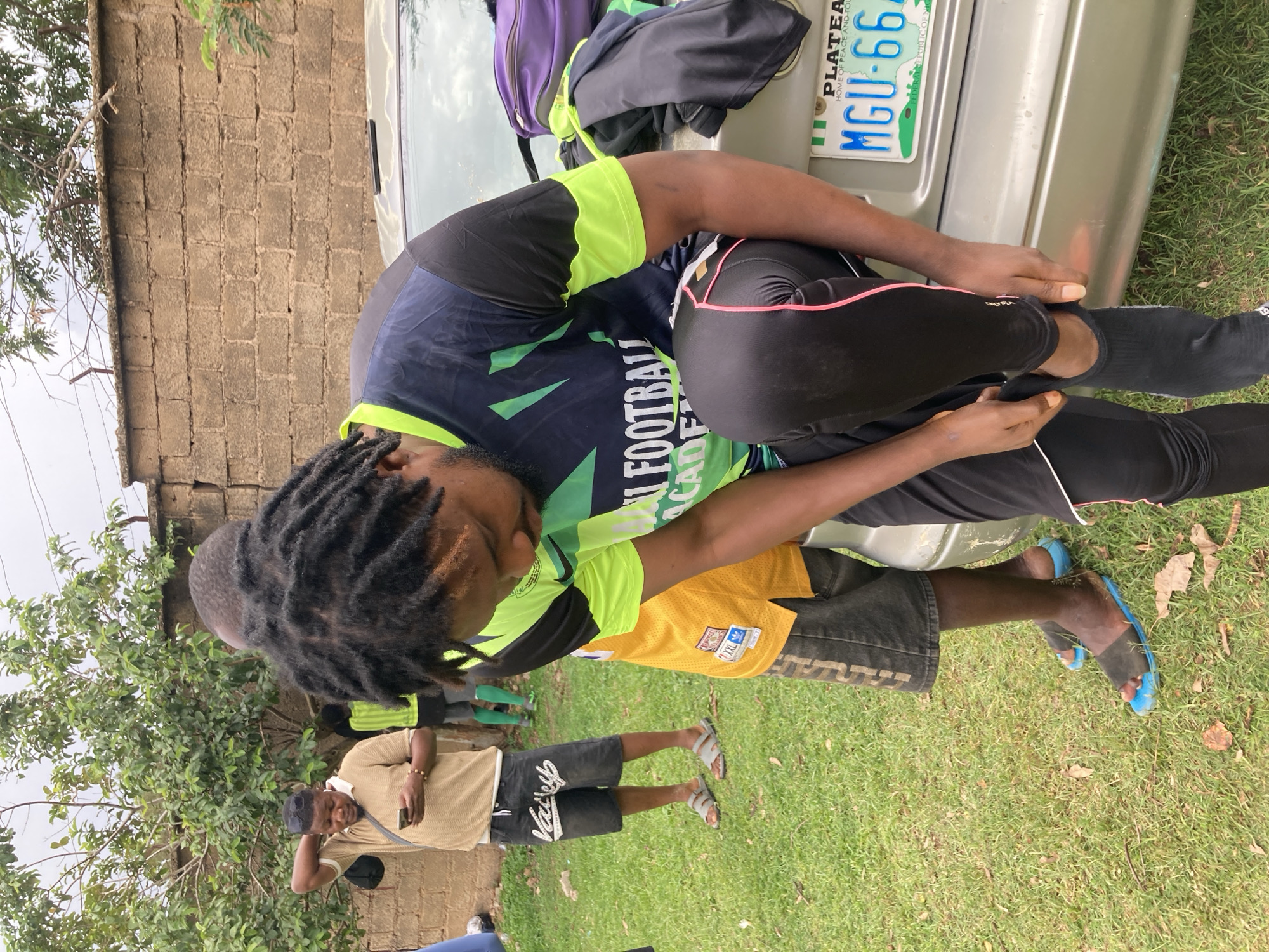 This outdoor image, taken in Gura Top, Nigeria, depicts a casual gathering of several individuals on a grassy area next to a silver car. The main subject, a person with dark dreadlocks, is seated on the car's bumper, engaged in adjusting black socks or footwear. This individual wears a black and neon green athletic shirt with "FOOTBAL ACADE" (likely "FOOTBALL ACADEMY") printed on it, paired with black athletic pants featuring a pink stripe.
In the background, a person in a light brown polo shirt and dark shorts, bearing the text "Val'ep XXL", stands casually, possibly holding a phone. Another person, partially visible, is wearing a yellow jersey, dark shorts, and blue slide sandals, standing on the grass.
The scene features a backdrop of a rough-hewn brick wall and green foliage from a large tree. The sky appears overcast, suggesting daytime. The rear of the silver car is visible, featuring a Nigerian license plate that reads "PLATEAU HOME OF PEACE AND TOURISM MGU-66 FEDERAL REPUBLIC OF NIGERIA," confirming the location context. Overall, the image conveys a relaxed atmosphere, possibly before or after an athletic event given the sports attire.