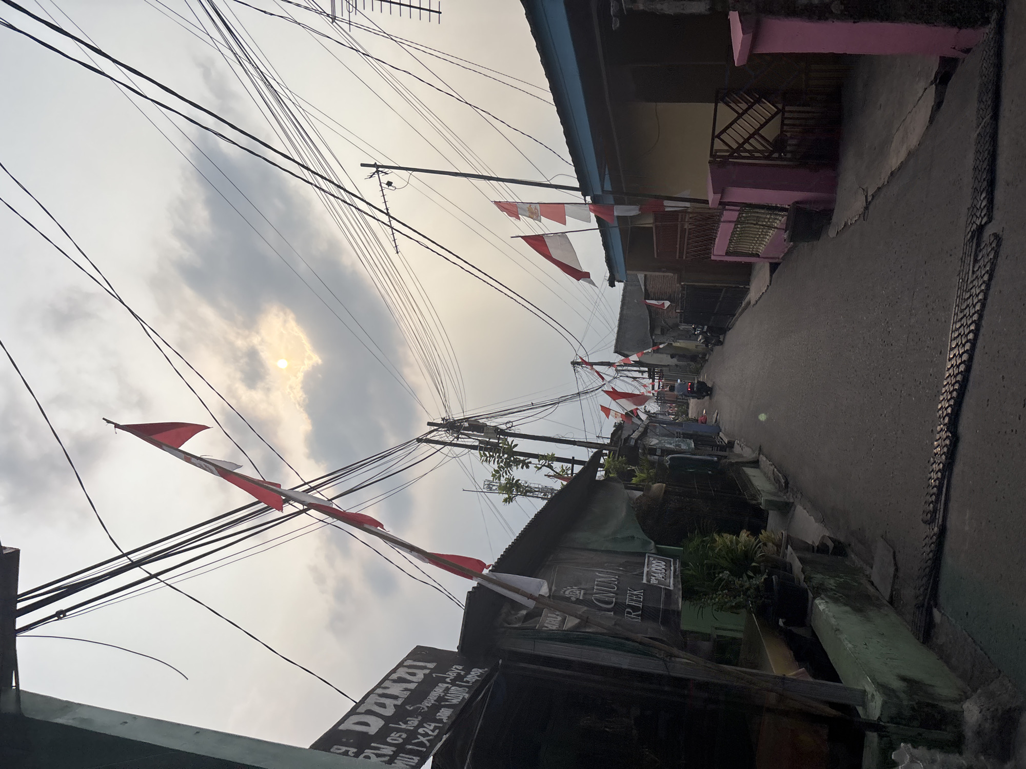 This is a photograph of a narrow, paved street in a residential area, likely in Bekasi, Indonesia, given the context. The street is lined with modest houses and small buildings. Indonesian flags, red and white, are prominently displayed on poles and draped from some of the buildings, suggesting a national holiday or celebration. The sky is overcast, with the sun peeking through the clouds, casting a soft light on the scene. Overhead, a dense network of electrical wires crisscrosses the sky, a common sight in many urban and semi-urban areas of Indonesia. On the left side of the street, a sign is visible on one of the buildings with text in Indonesian. A motorcycle is seen in the distance on the right side of the street, with a rider, and another motorcycle is parked on the left. The overall impression is one of a bustling, lived-in community.