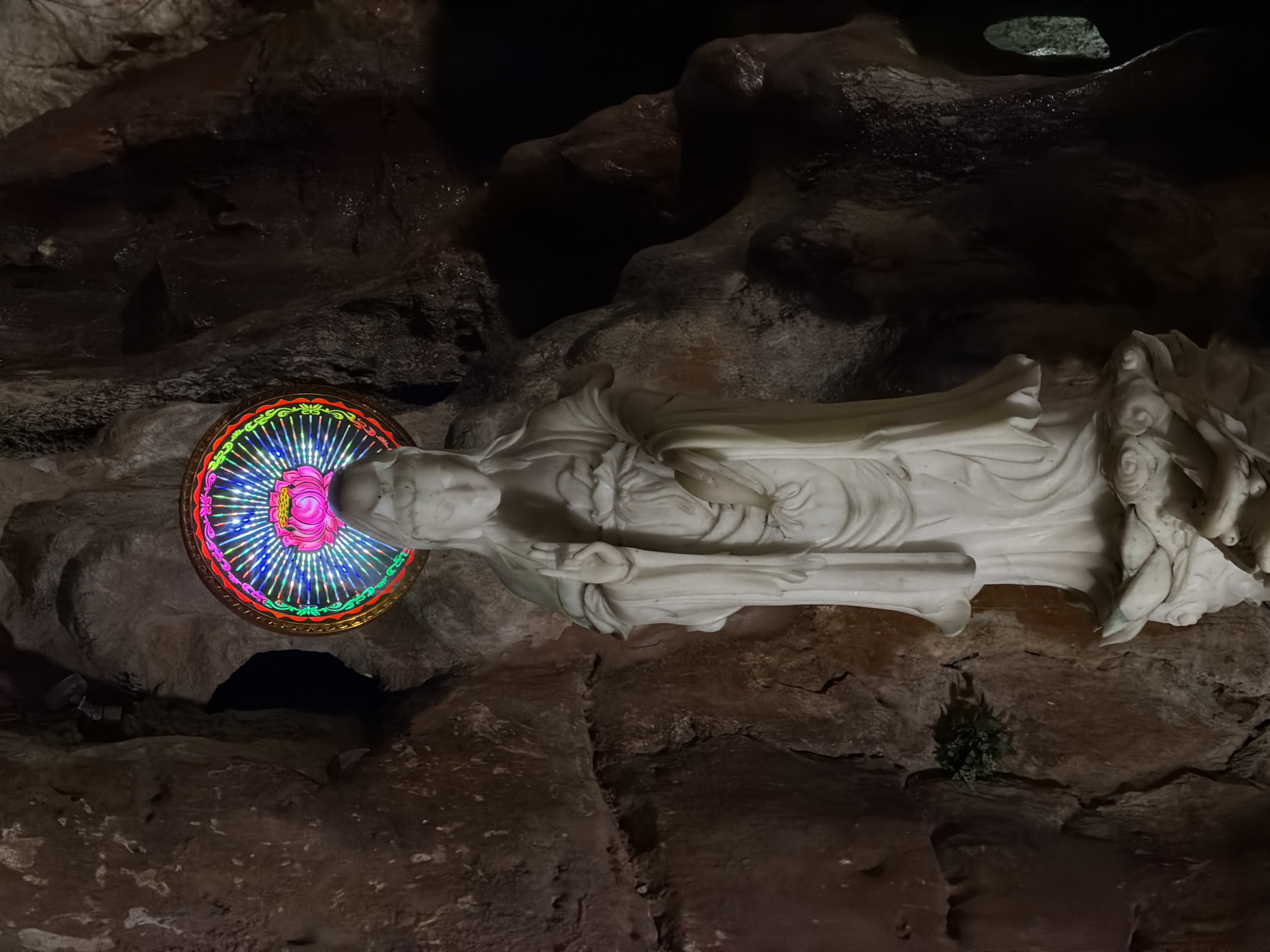 A statue of a woman, illuminated by a circular light, is positioned within a cavern. The statue is white, and the light surrounding its head is multicolored, featuring a pink center. The cavern is composed of dark brown rock formations. The statue is located in Đà Nẵng, Vietnam.