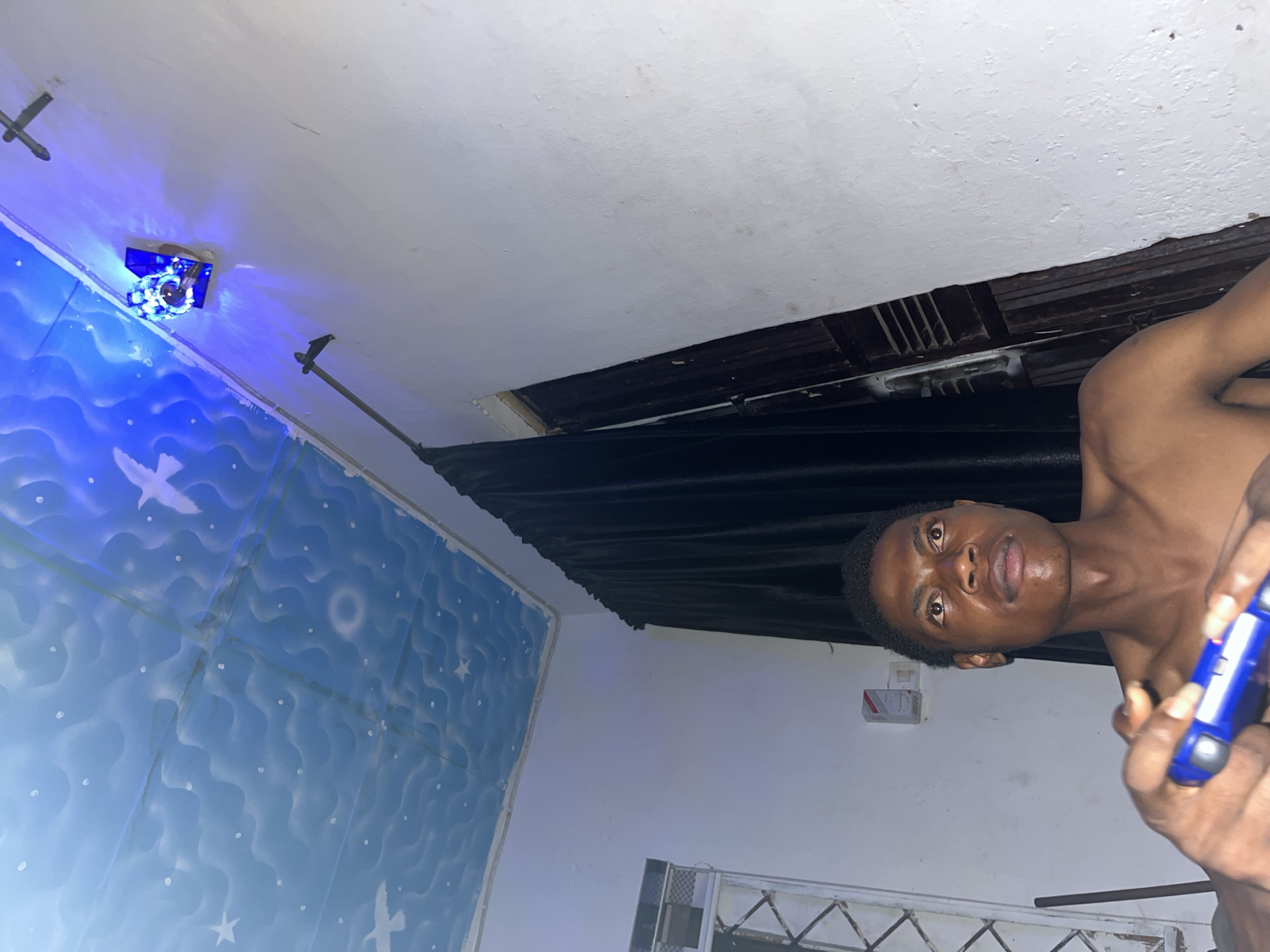 An indoor scene captured with the camera rotated 90 degrees clockwise features a young man with dark skin and short dark hair, looking directly at the viewer with a neutral expression. He is shirtless and holds a blue game controller in both hands, with his fingers positioned on the buttons.
The background reveals a mix of finished and unfinished elements. The ceiling is white, but a prominent section shows exposed wooden beams or joists, suggesting an unfinished construction or damage. Below this, a dark, thick curtain is drawn. On the left side of the image, a wall is decorated with multiple blue panels featuring white wave patterns, stars, and silhouettes of birds, giving an ethereal or aquatic theme. A blue light fixture, possibly decorative, emits a bright blue glow onto these panels. Further behind the man, on a plain white wall, a small white electrical box with red markings is visible. In the lower right corner, a section of a metal gate or railing can be seen. The overall lighting suggests an interior space, likely in the evening or night, relying on artificial light sources. No discernible text is visible.